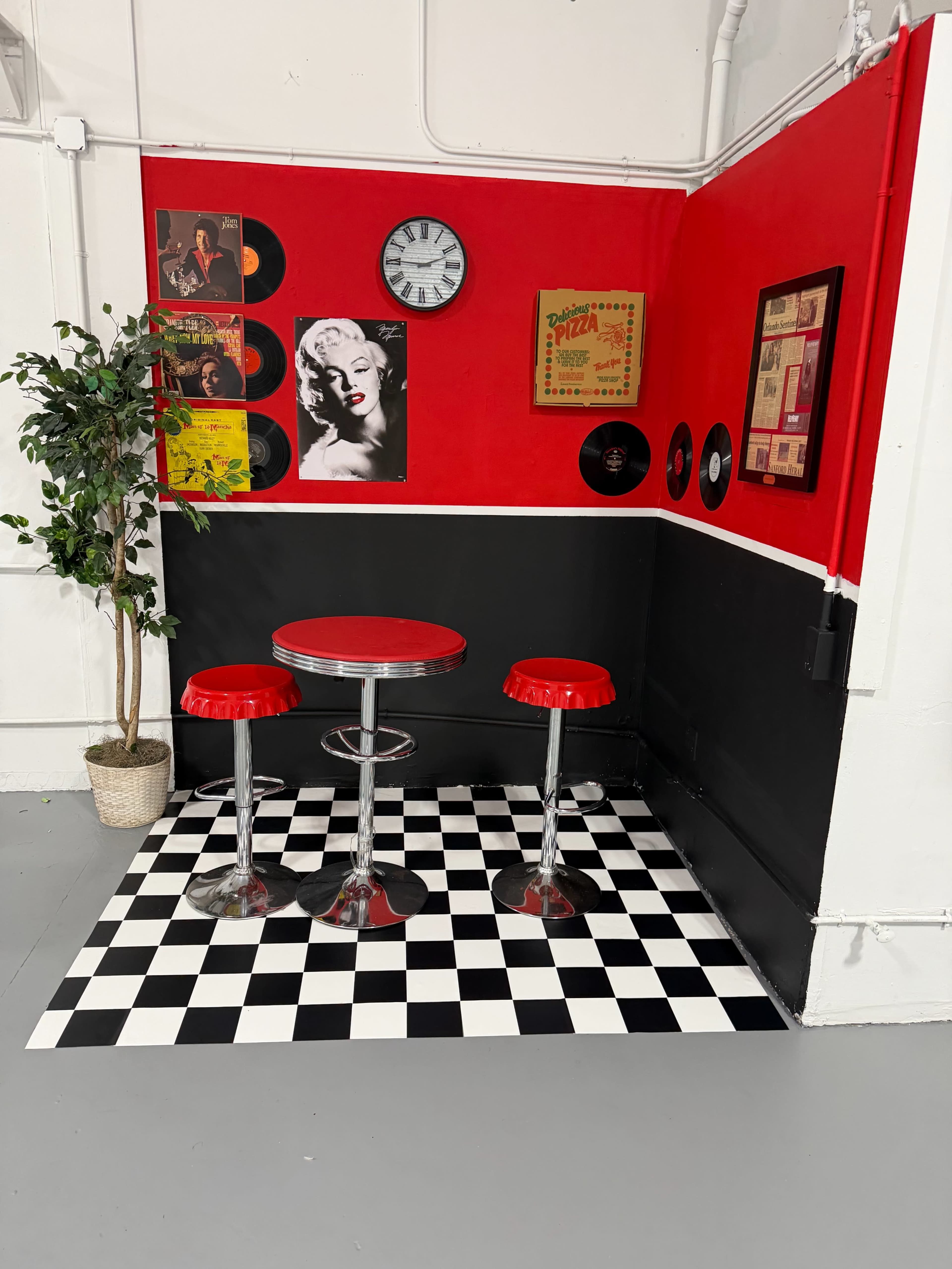 The image depicts a retro-themed diner corner with a red and black color scheme, featuring a round table with two stools, a checkered floor, and vintage decor on the walls.