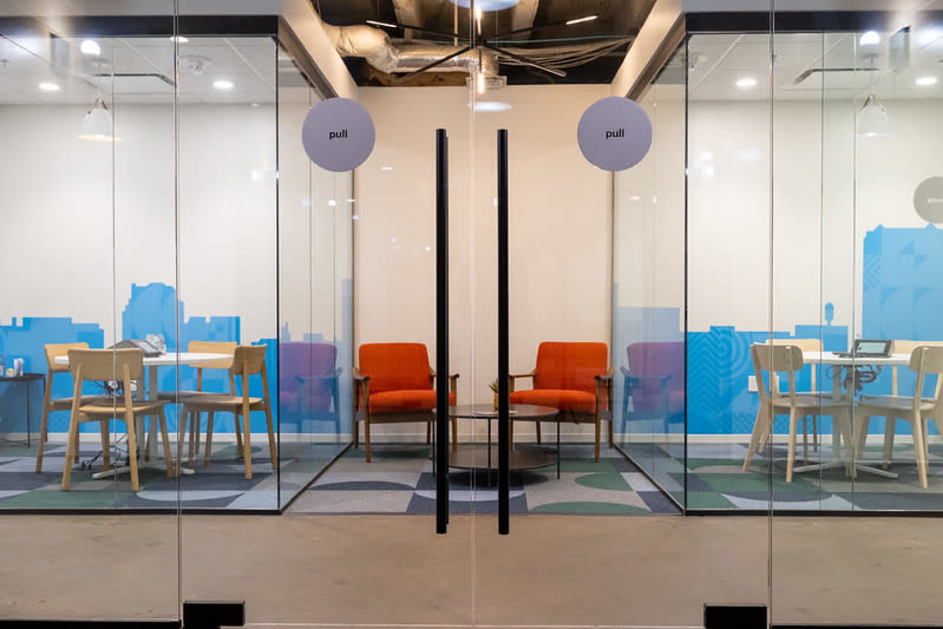 The image shows a glass-walled office space with two meeting areas, each featuring a table and chairs, separated by a central door marked "pull."