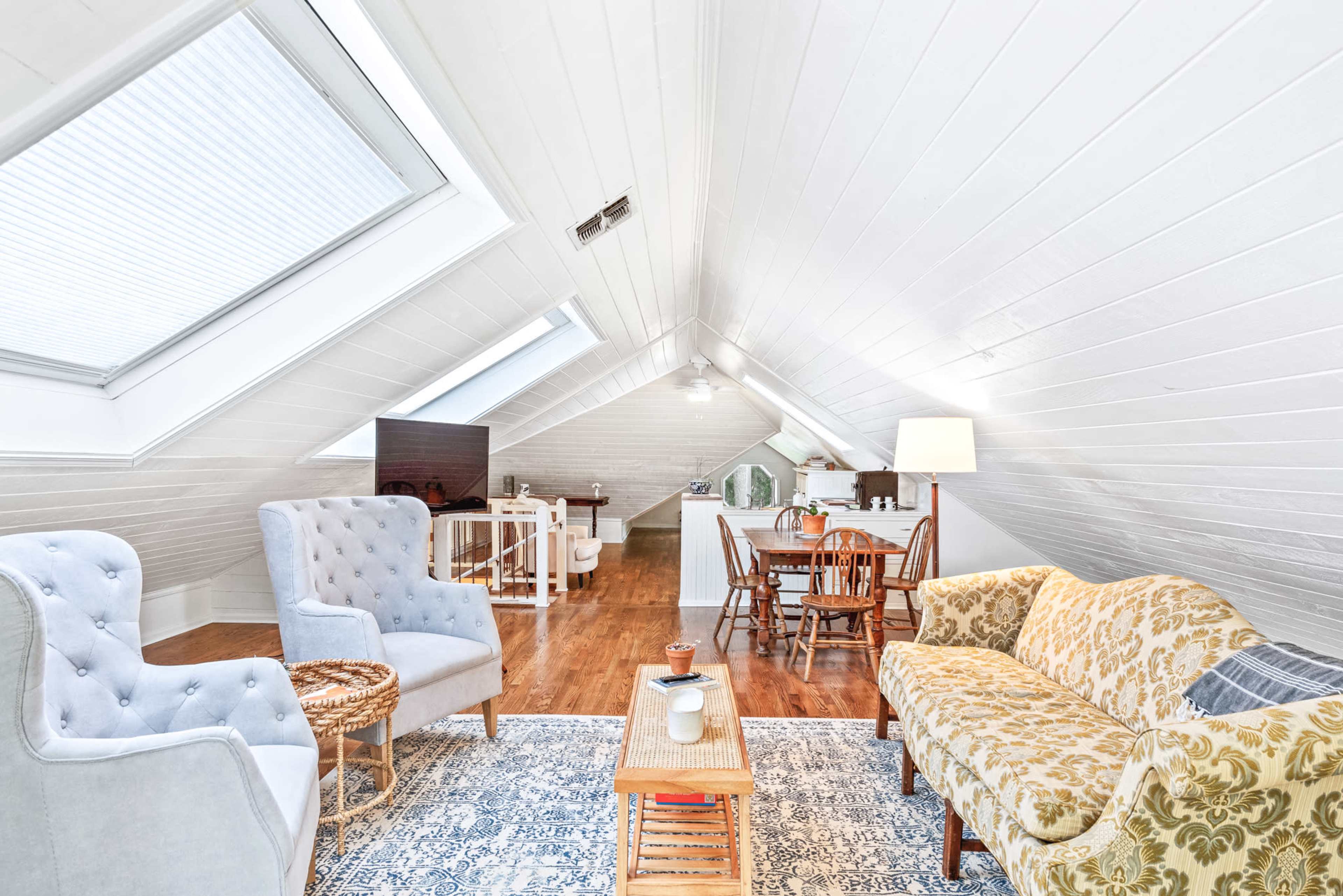 An airy attic space features a cozy seating area with two armchairs and a patterned sofa, alongside a dining table and kitchenette.