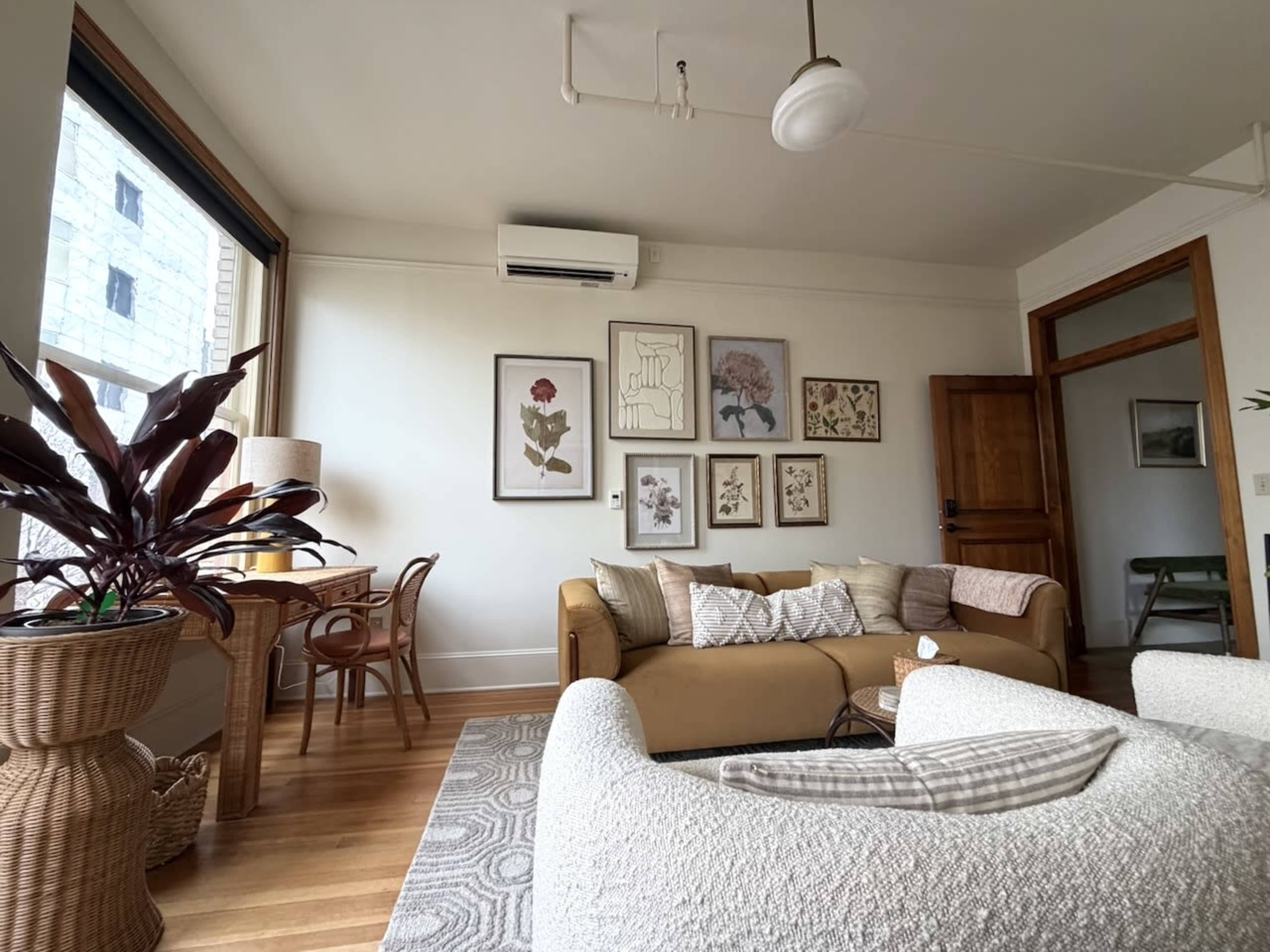 The image shows a cozy living room featuring a tan sofa, a white armchair, a desk with a lamp, and framed botanical art on the walls.
