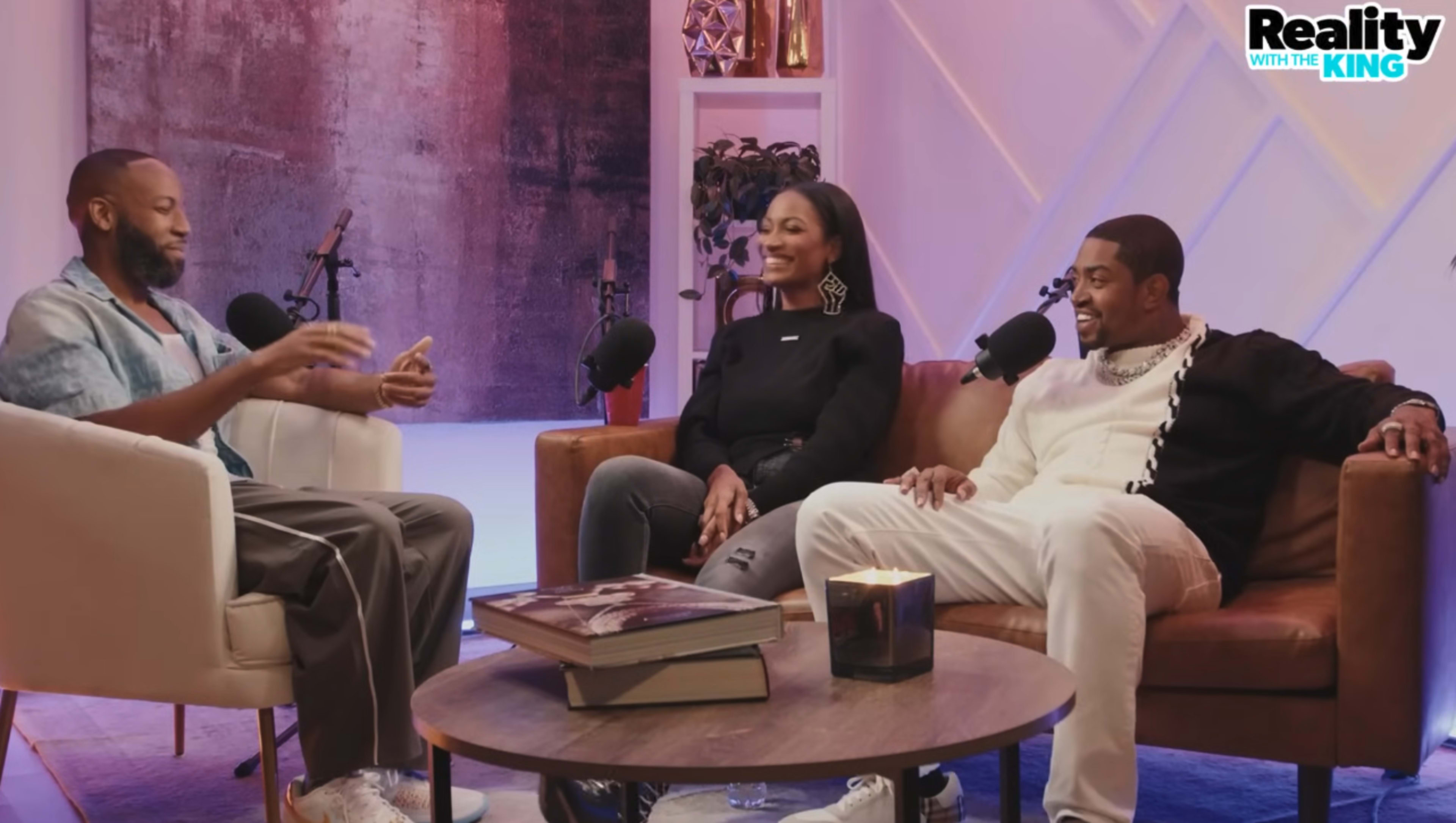 Three individuals are seated in a modern studio environment discussing something animatedly, with a coffee table in front of them that holds a book and a candle.