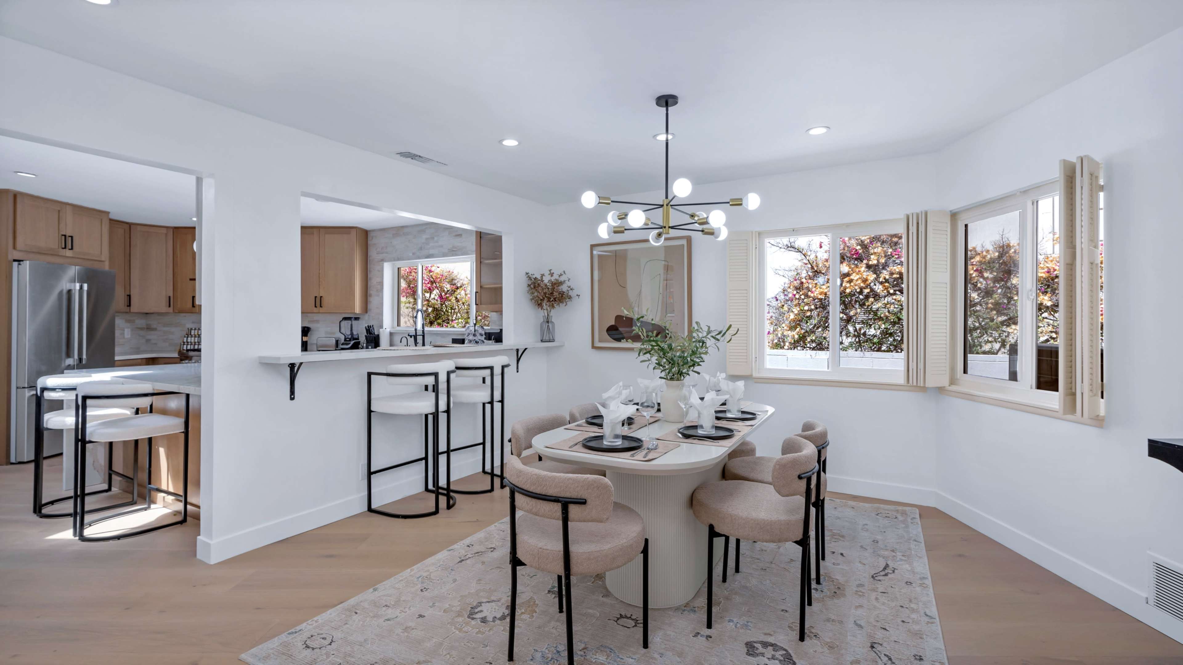 A modern dining area features a round table surrounded by upholstered chairs, with a view of trees visible through large windows.