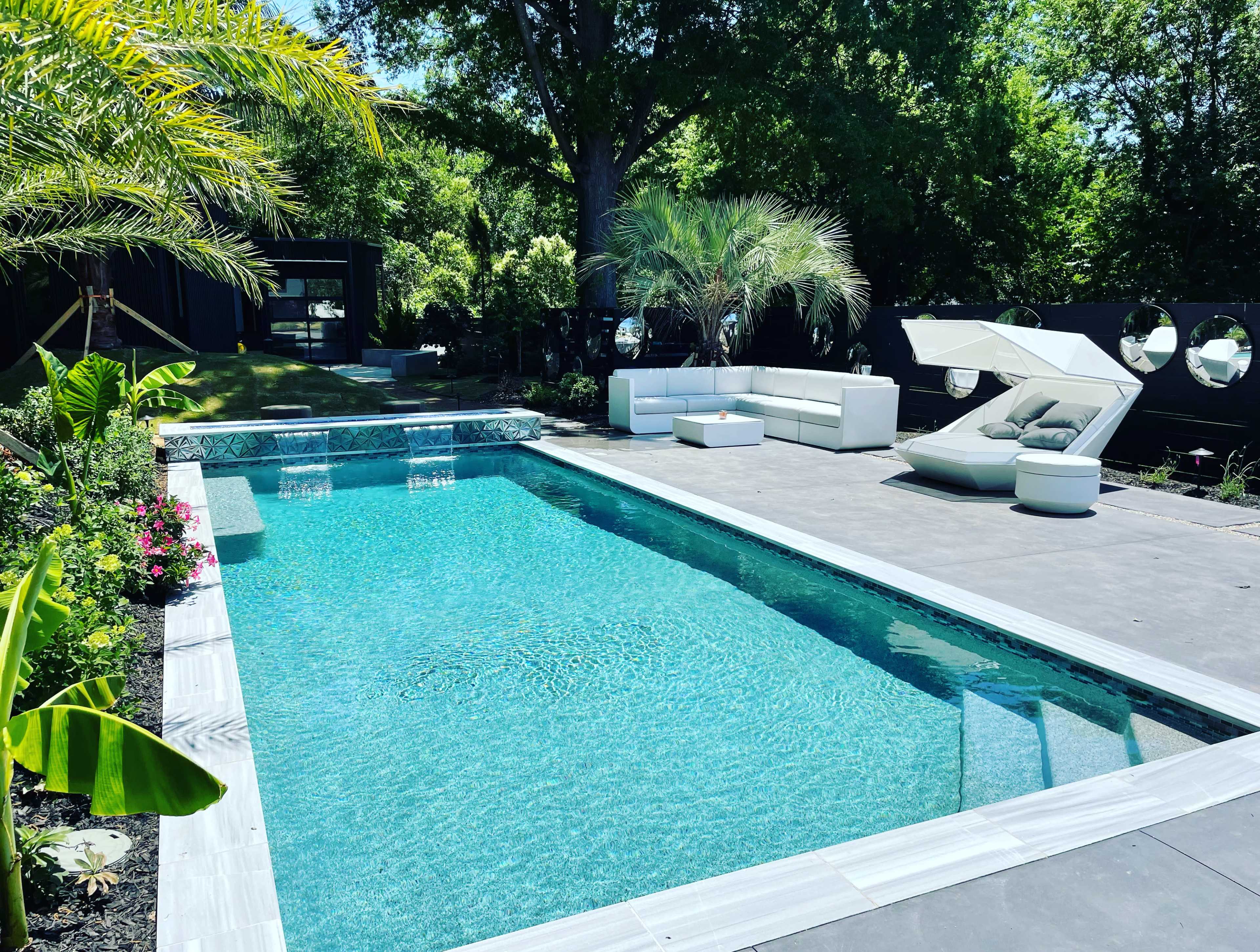 The image shows a modern swimming pool surrounded by landscaped greenery, featuring lounge seating and a sleek sunshade.