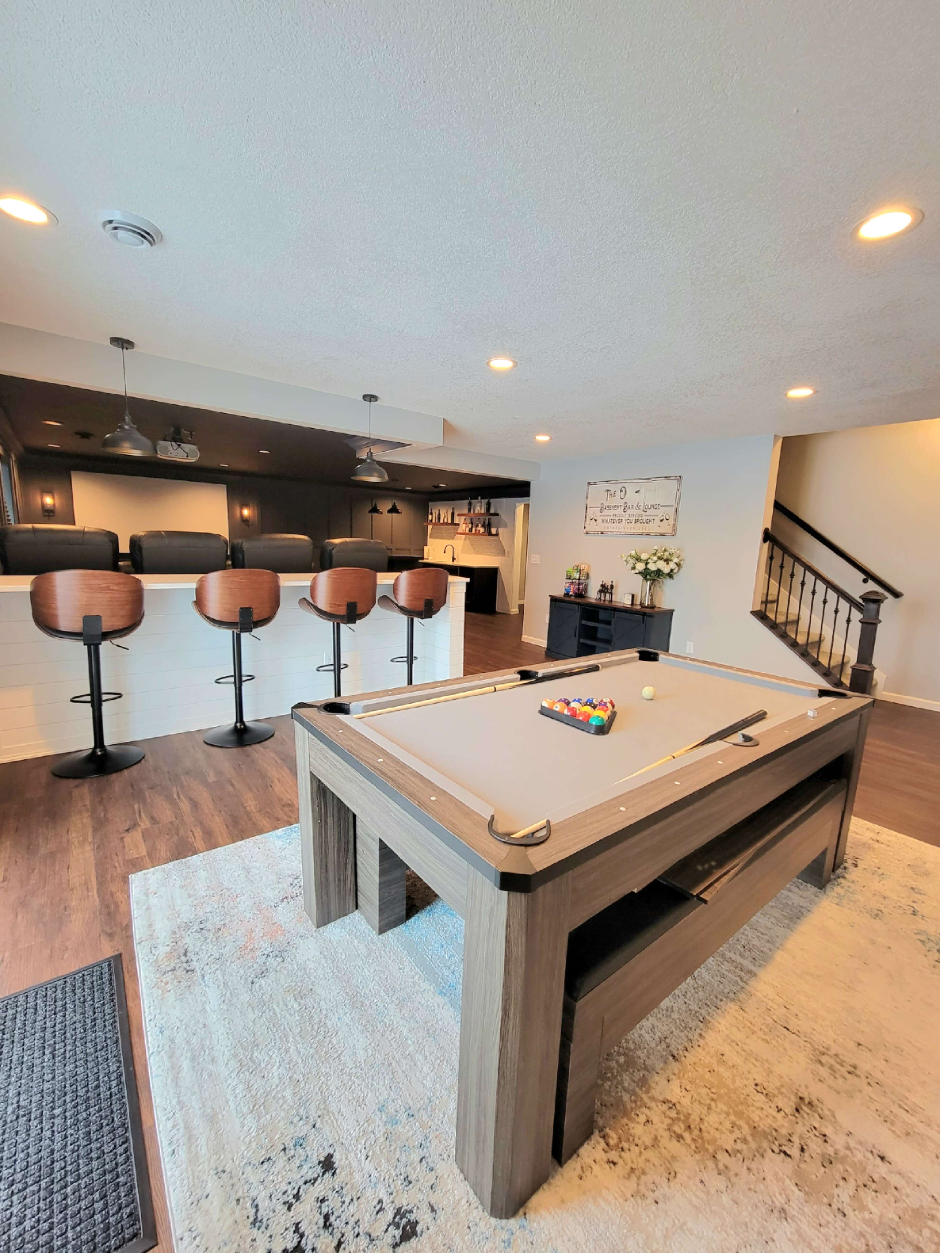 A modern game room featuring a pool table at the center, surrounded by a seating area with bar stools and a wet bar in the background.