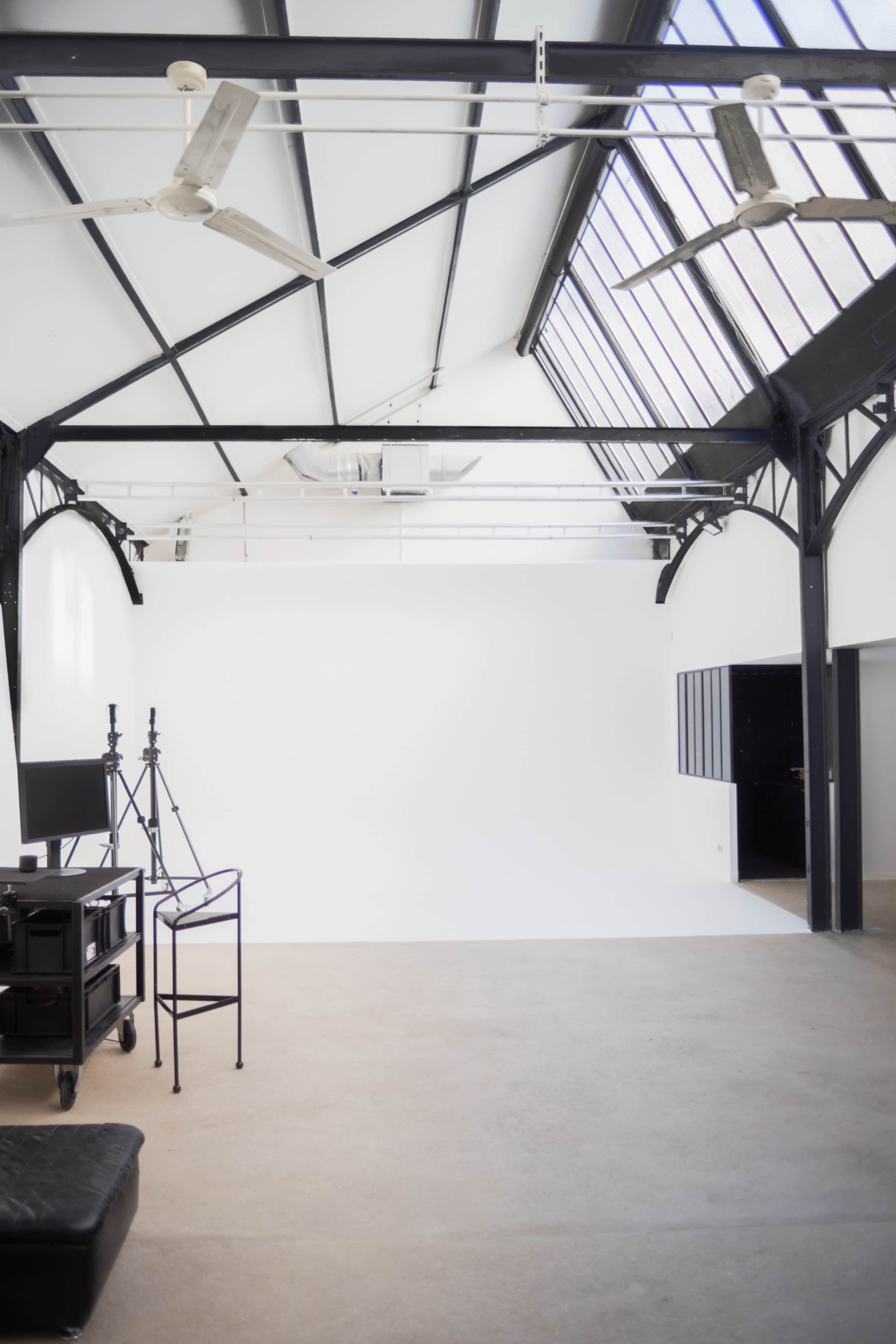 The image shows a spacious, modern studio with a high ceiling, large windows, and a plain white backdrop.