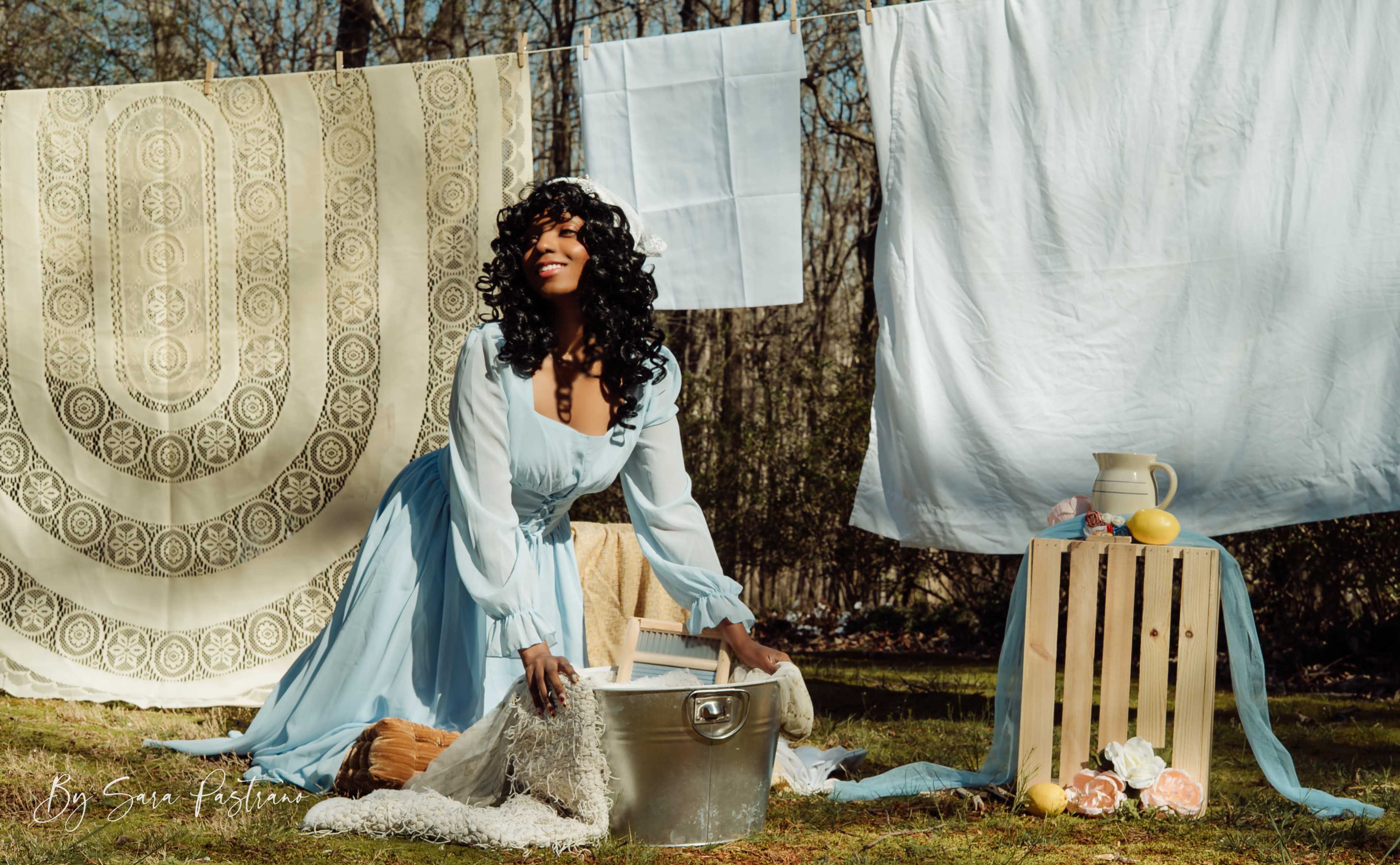 A woman in a light blue dress kneels beside a metal basin outdoors, surrounded by hanging fabric and a wooden crate with decorative items.