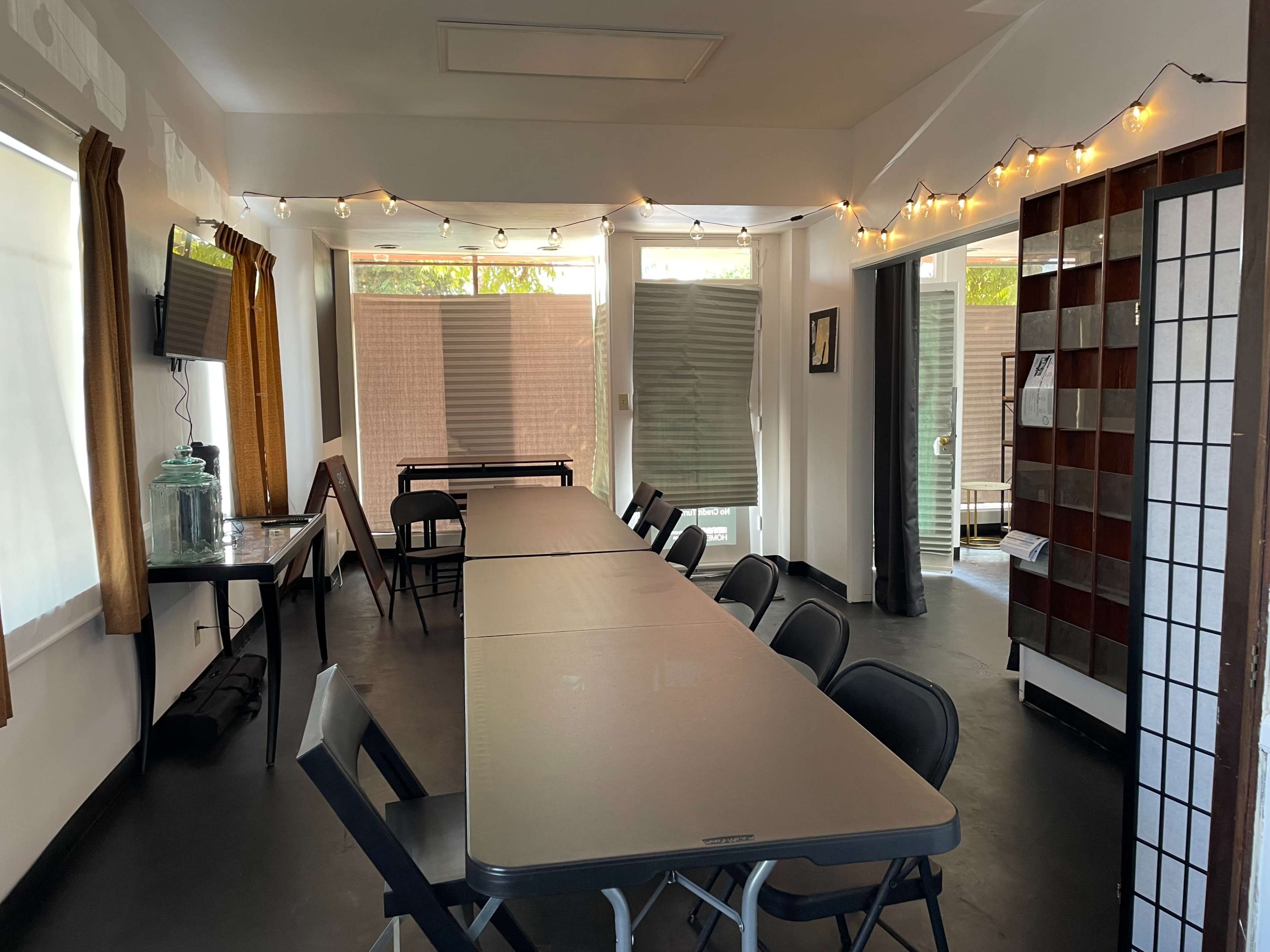 A room with a long table set up with folding chairs, string lights along the walls, and large windows partially covered by blinds.