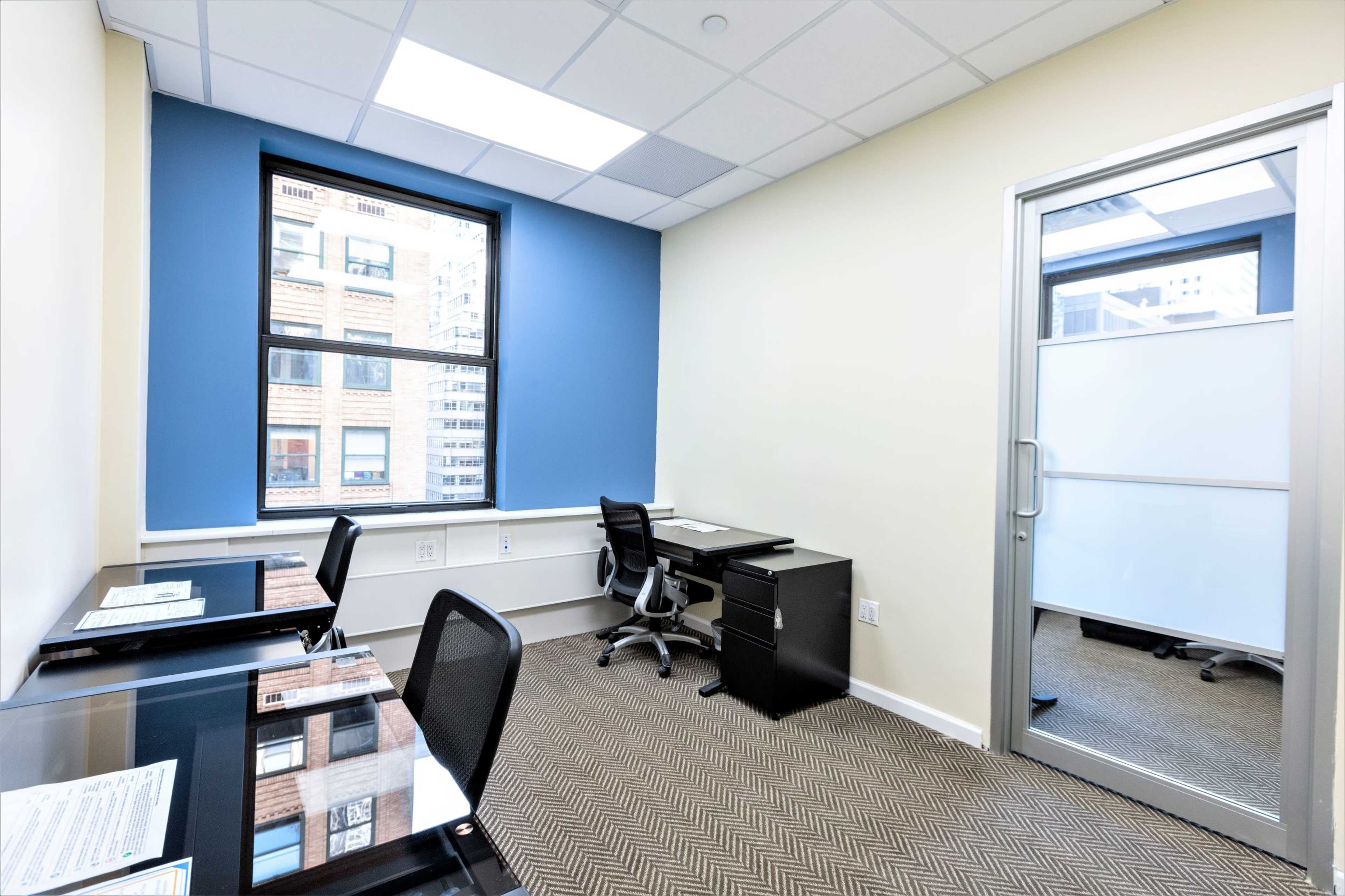 The image shows a small office space with two desk setups, a black office chair, and a large window letting in natural light.