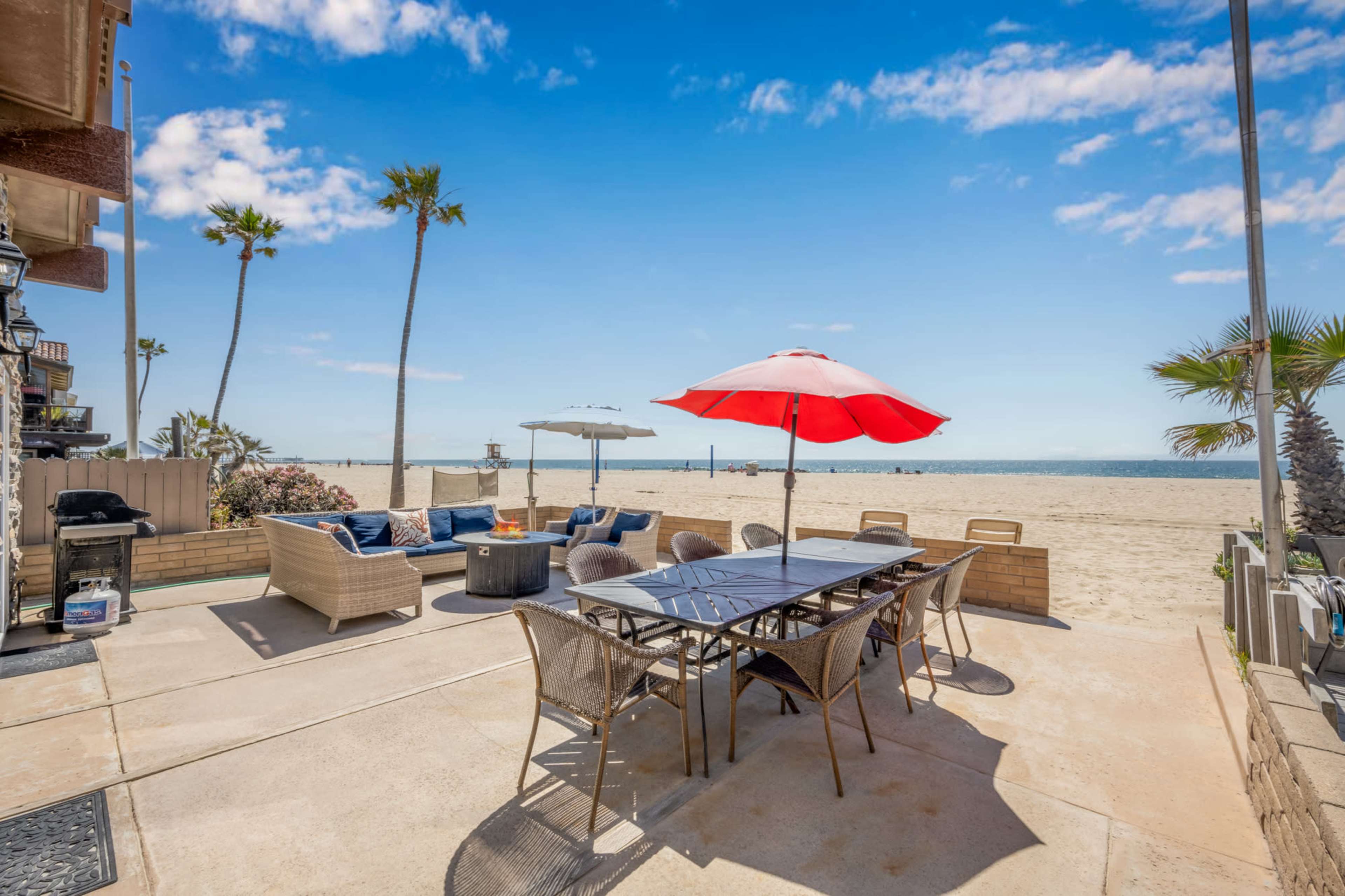 A patio area with a table and umbrellas overlooks a sandy beach and ocean under a clear blue sky.
