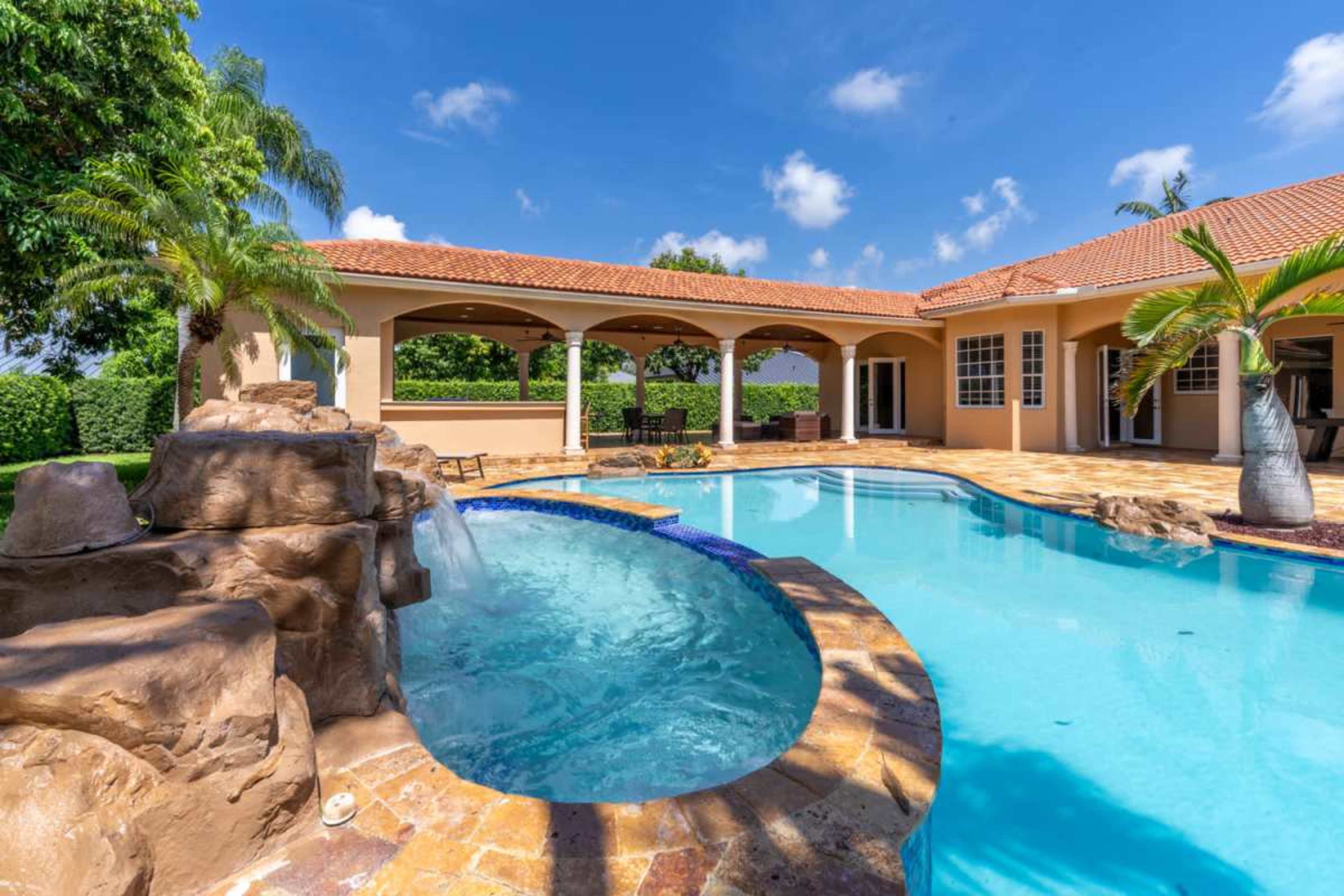 The image shows a backyard pool area featuring a curved swimming pool, a spa with a waterfall, and a house with an arched patio and tropical landscaping.