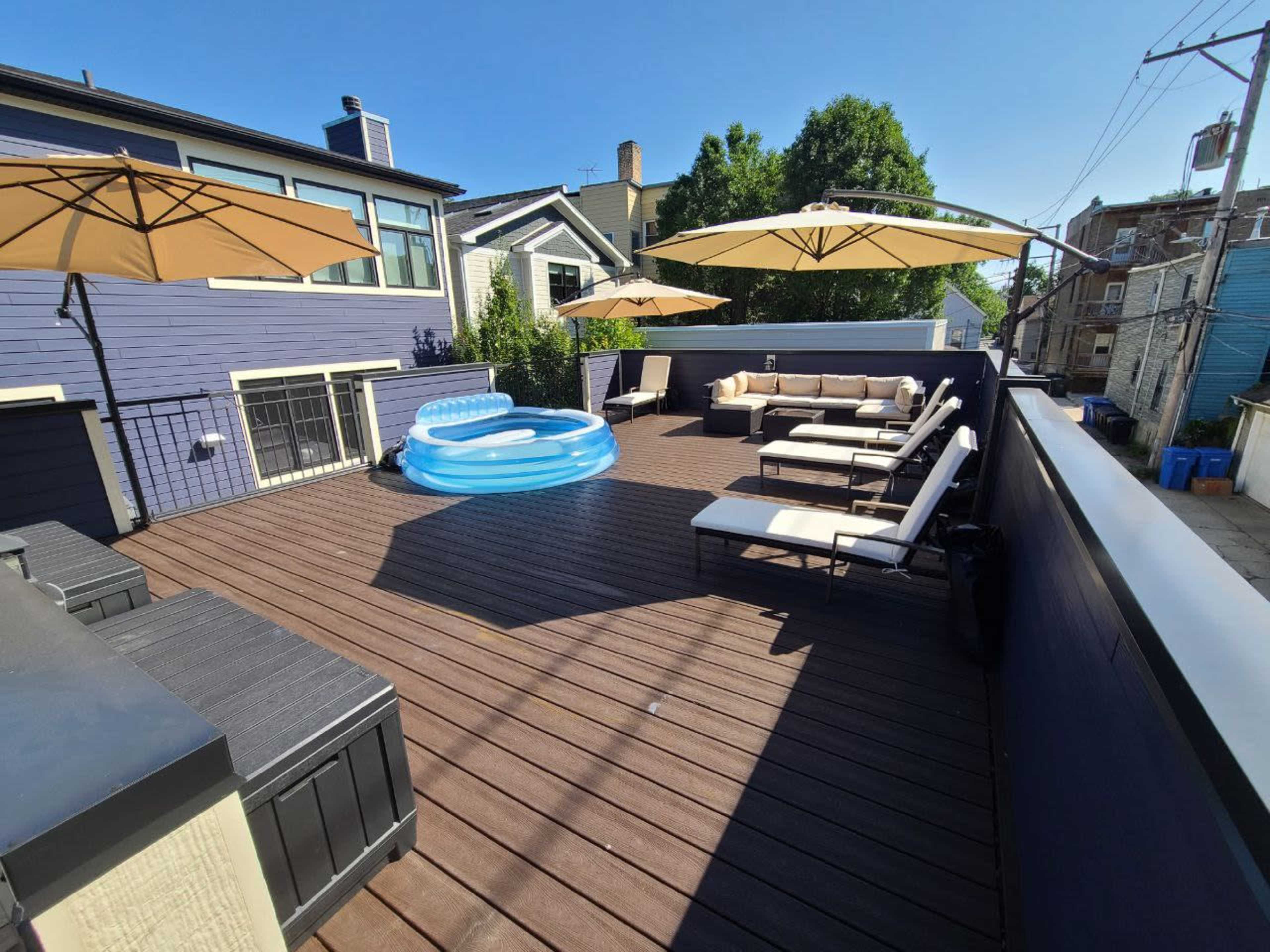 A spacious wooden deck equipped with lounge chairs, a beige sectional sofa, a circular inflatable pool, and several umbrellas for shade.