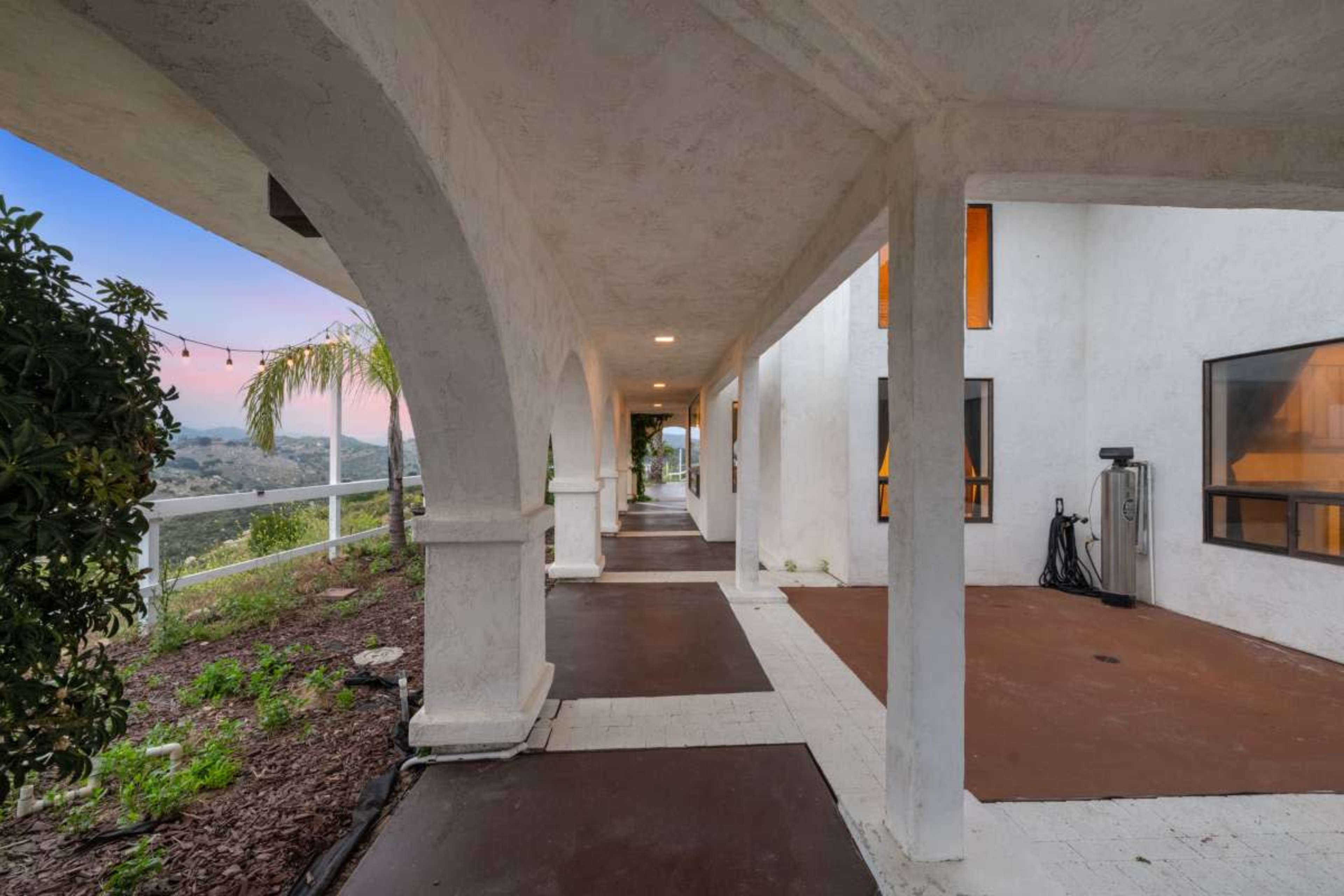 A covered walkway lined with columns leads to a series of windows overlooking a hillside landscape.