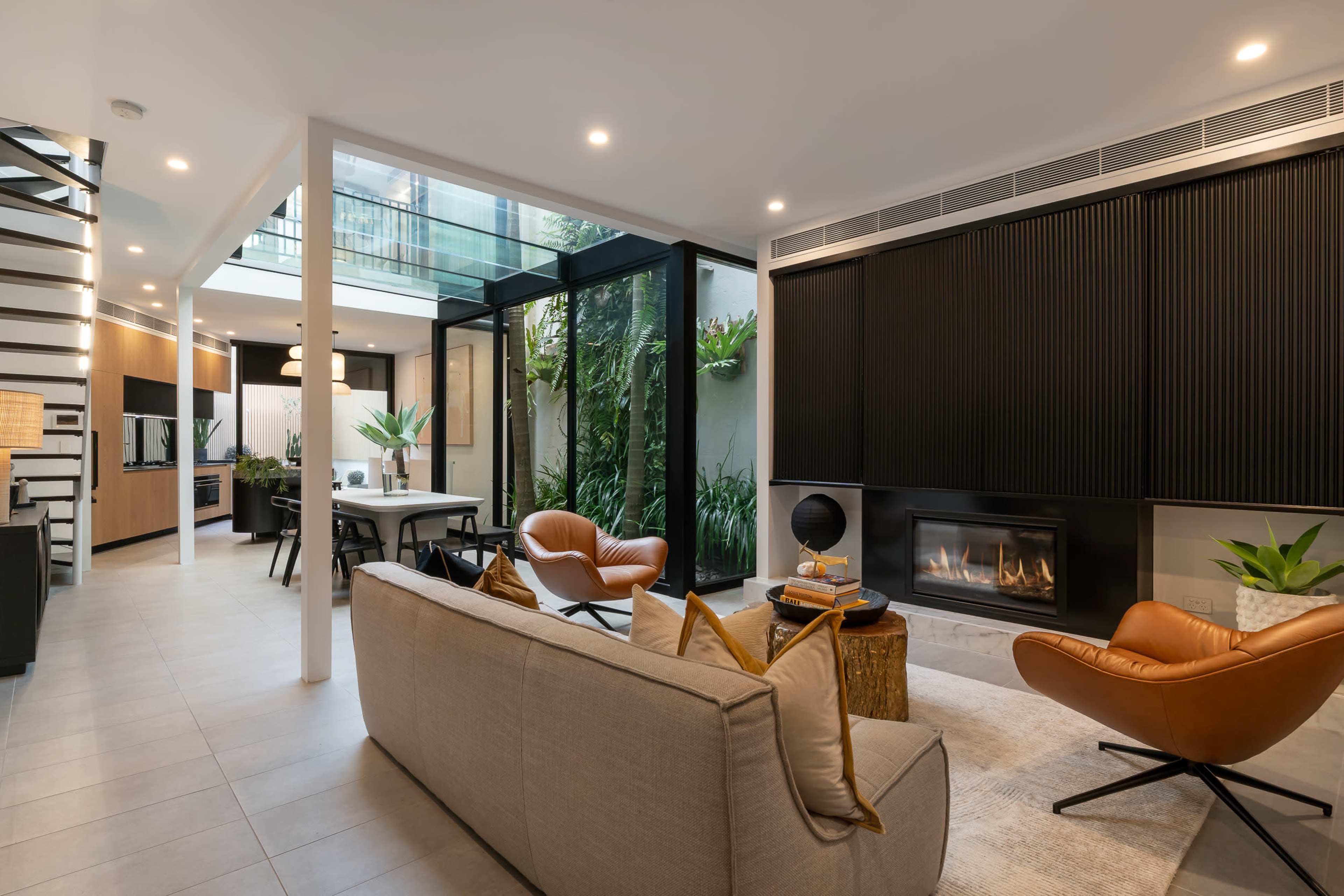 The image shows a modern living space with a sleek design, featuring a living area with brown chairs, a fireplace, and a view of an indoor garden.