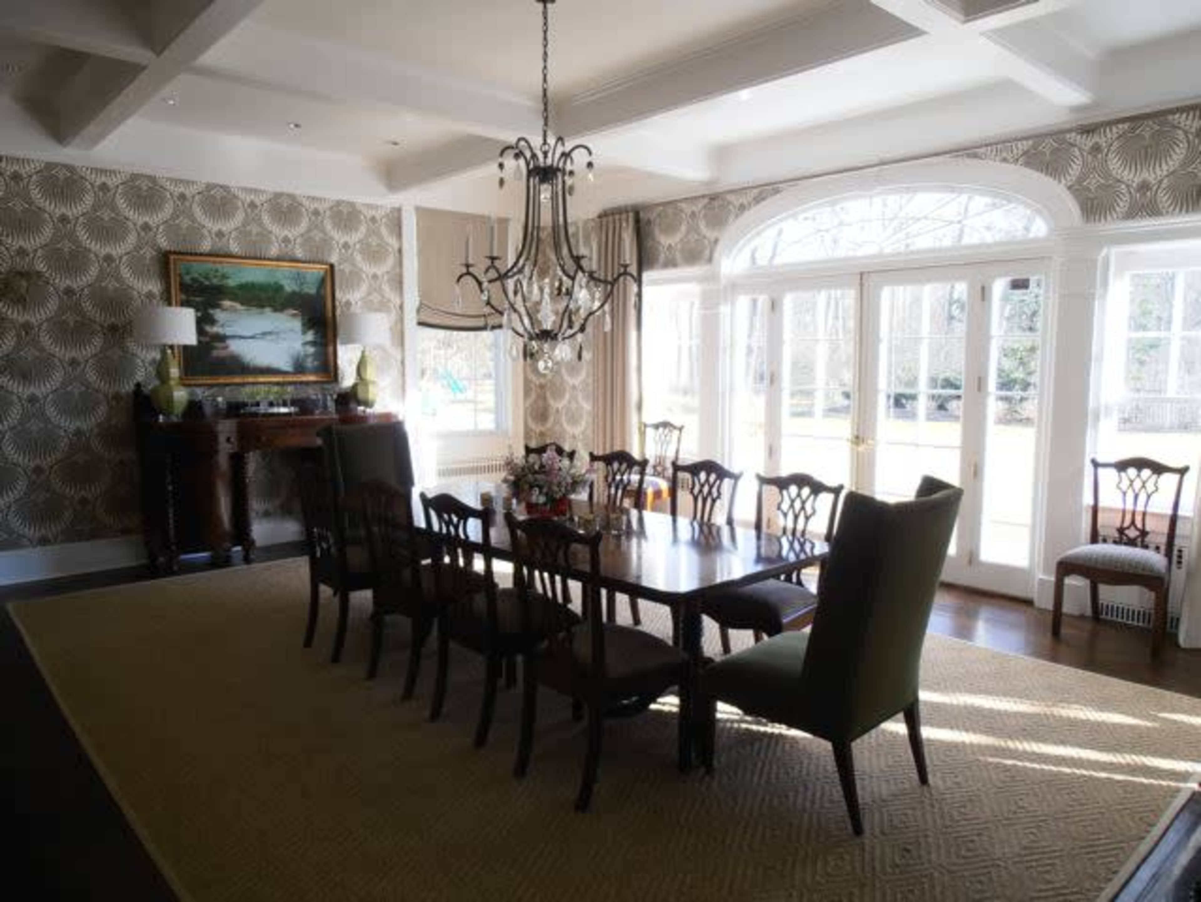 A spacious dining room features a long wooden table surrounded by chairs, a chandelier overhead, and large windows allowing natural light to enter.
