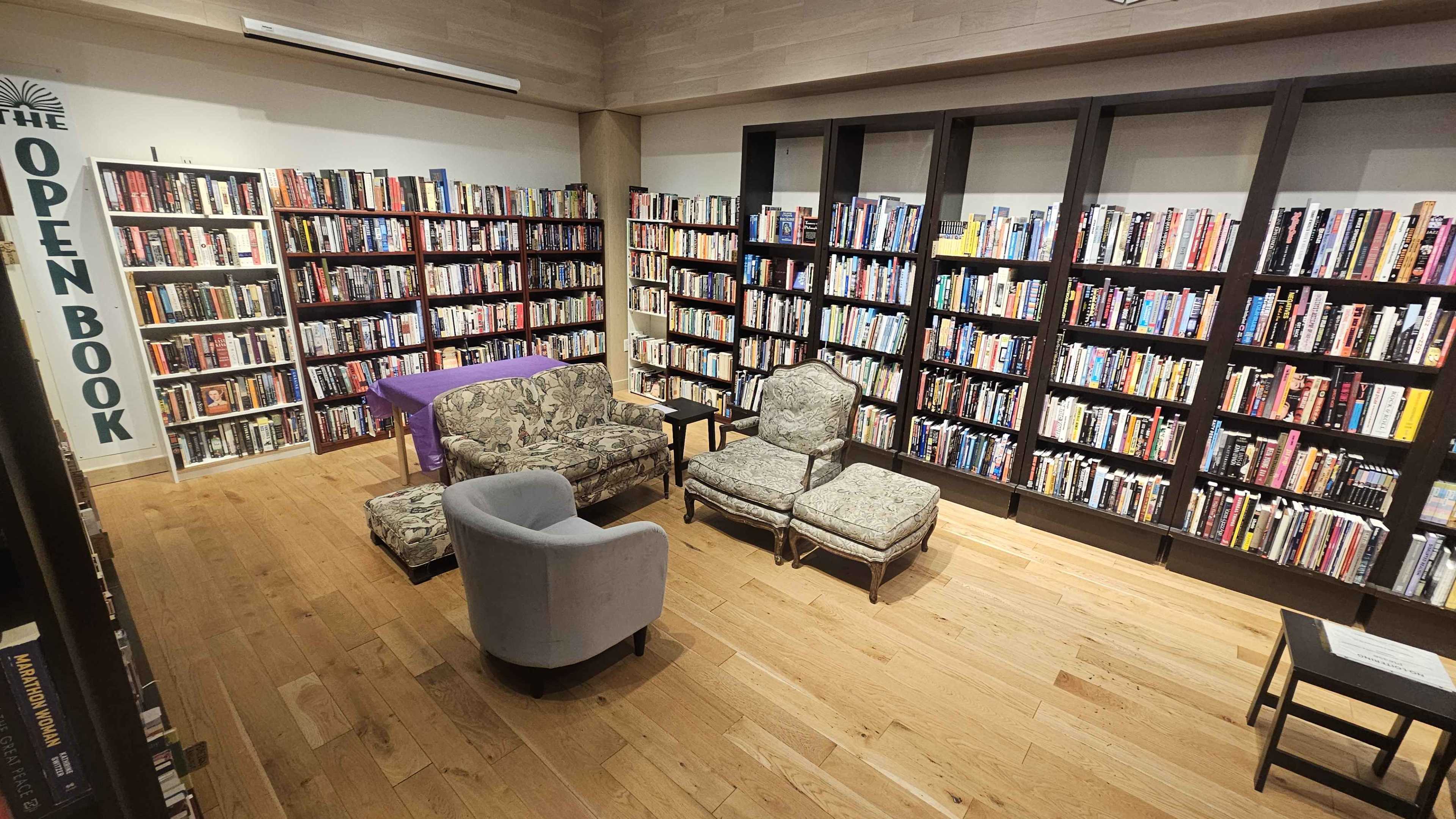 The image shows a cozy library space with bookshelves filled with various books, two patterned armchairs, a light blue chair, and a small table.