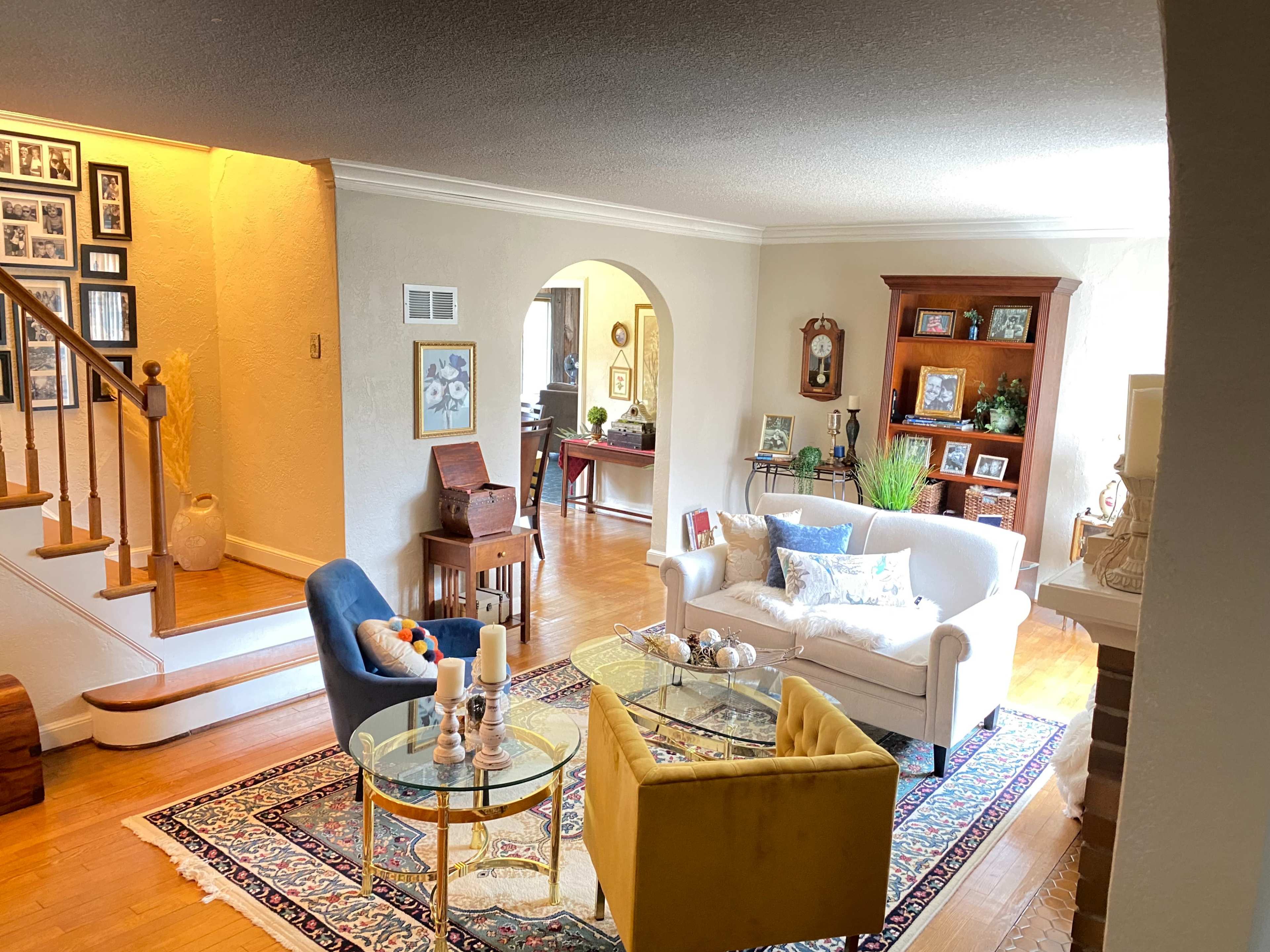 A living room features a white sofa and yellow chairs arranged around a glass coffee table, with a staircase in the background and framed photographs on the wall.