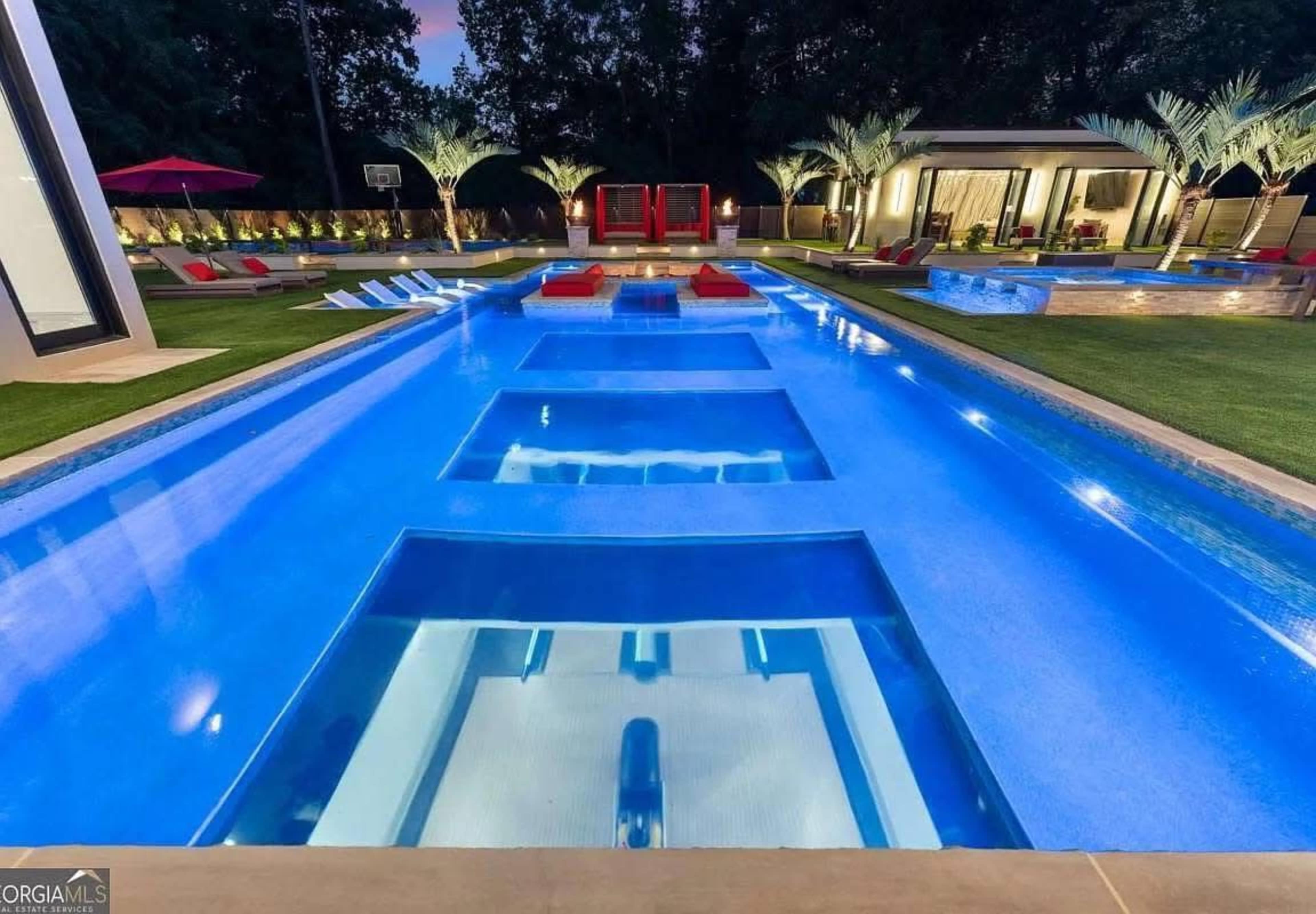 The image shows a modern swimming pool area featuring multiple water channels, lounge chairs, and palm trees, illuminated by soft outdoor lighting at dusk.