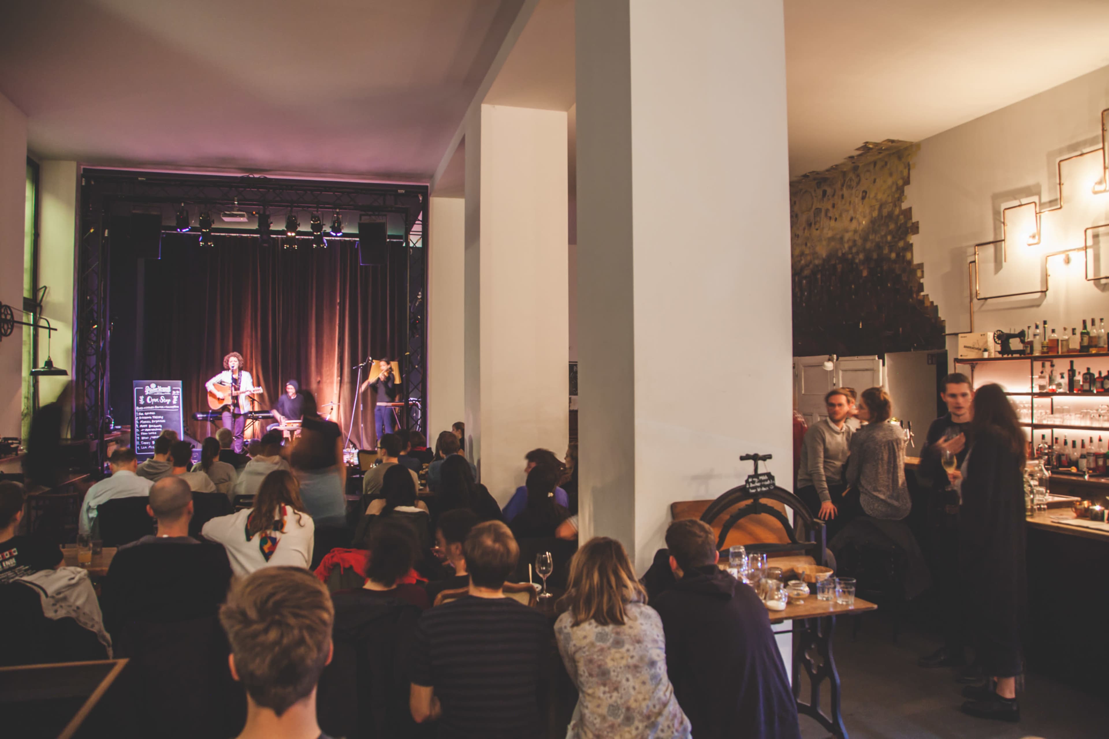 A performer plays guitar on stage in a dimly lit venue filled with an audience seated at tables and standing around the bar.
