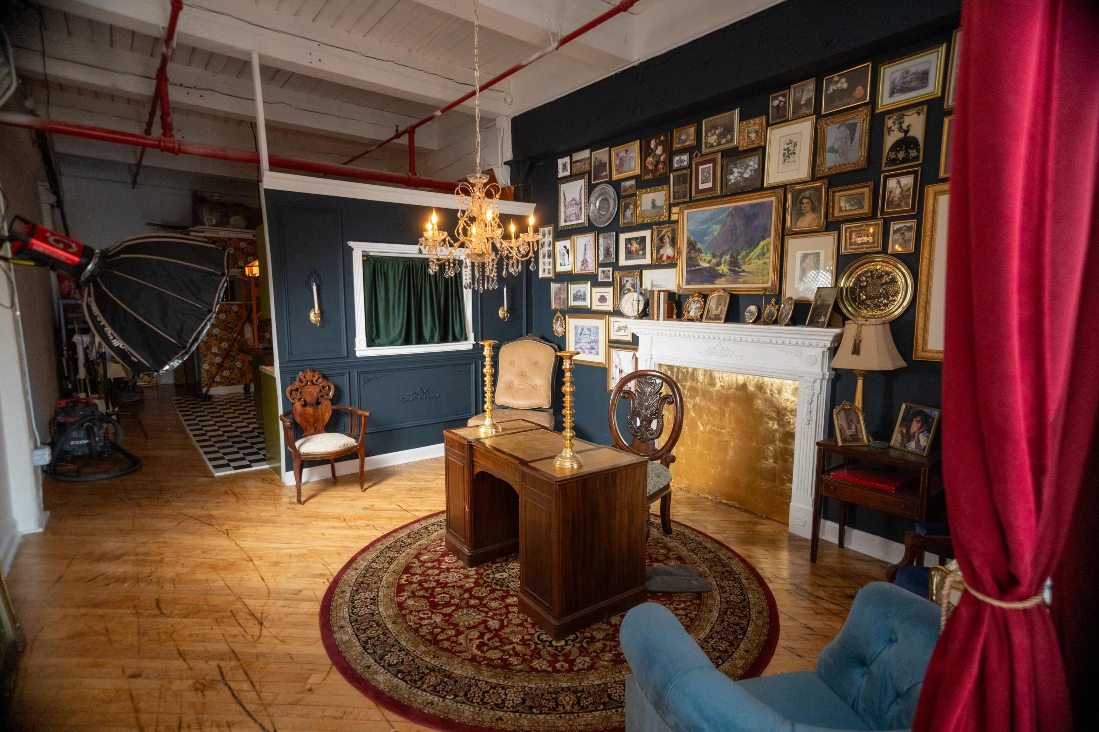 The room features a vintage wooden desk centered on a circular rug, surrounded by framed art on the dark blue walls and a fireplace with a golden surround.