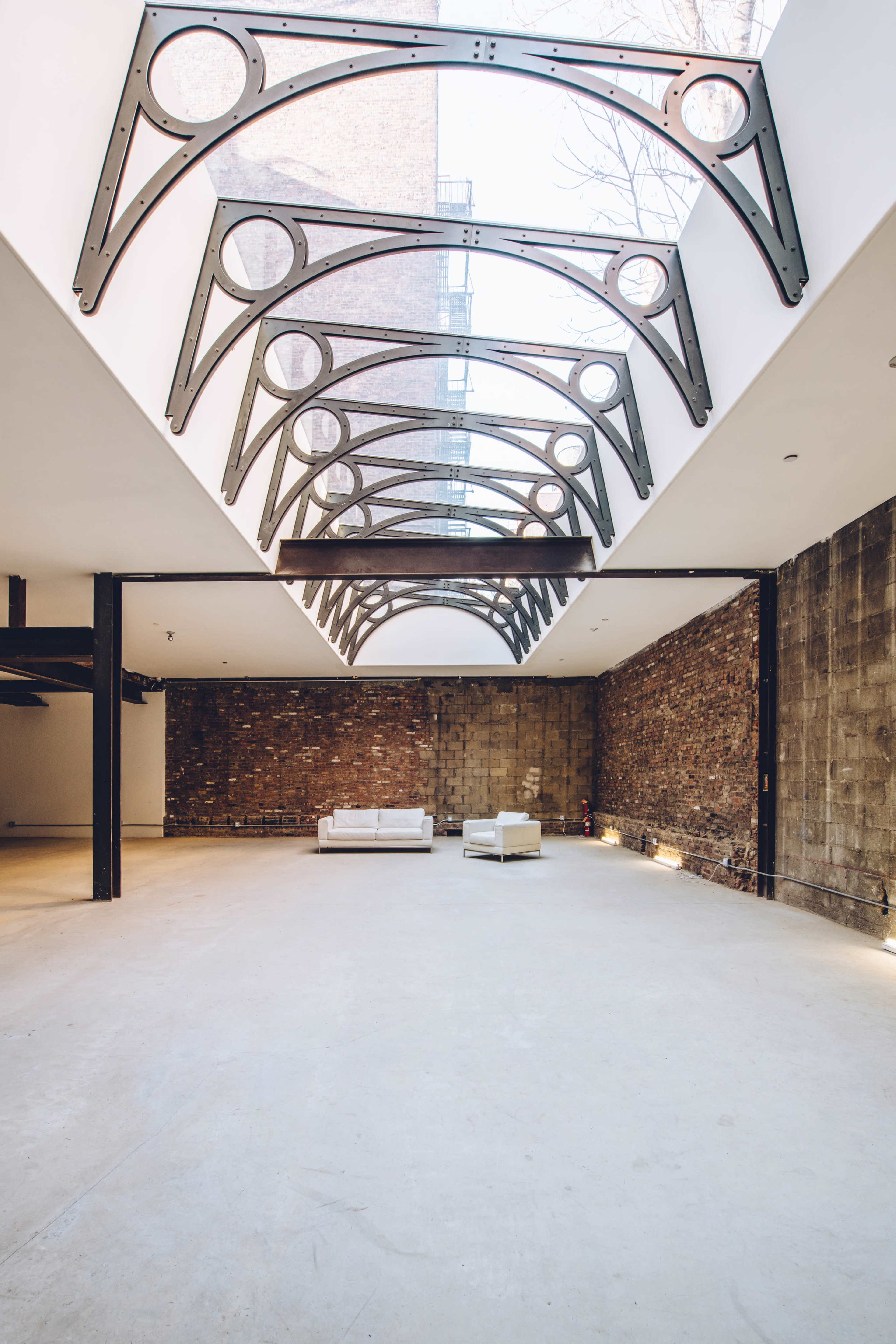 The image shows a spacious, minimalist interior with exposed brick walls, a concrete floor, and a ceiling featuring large metal arches and skylights.