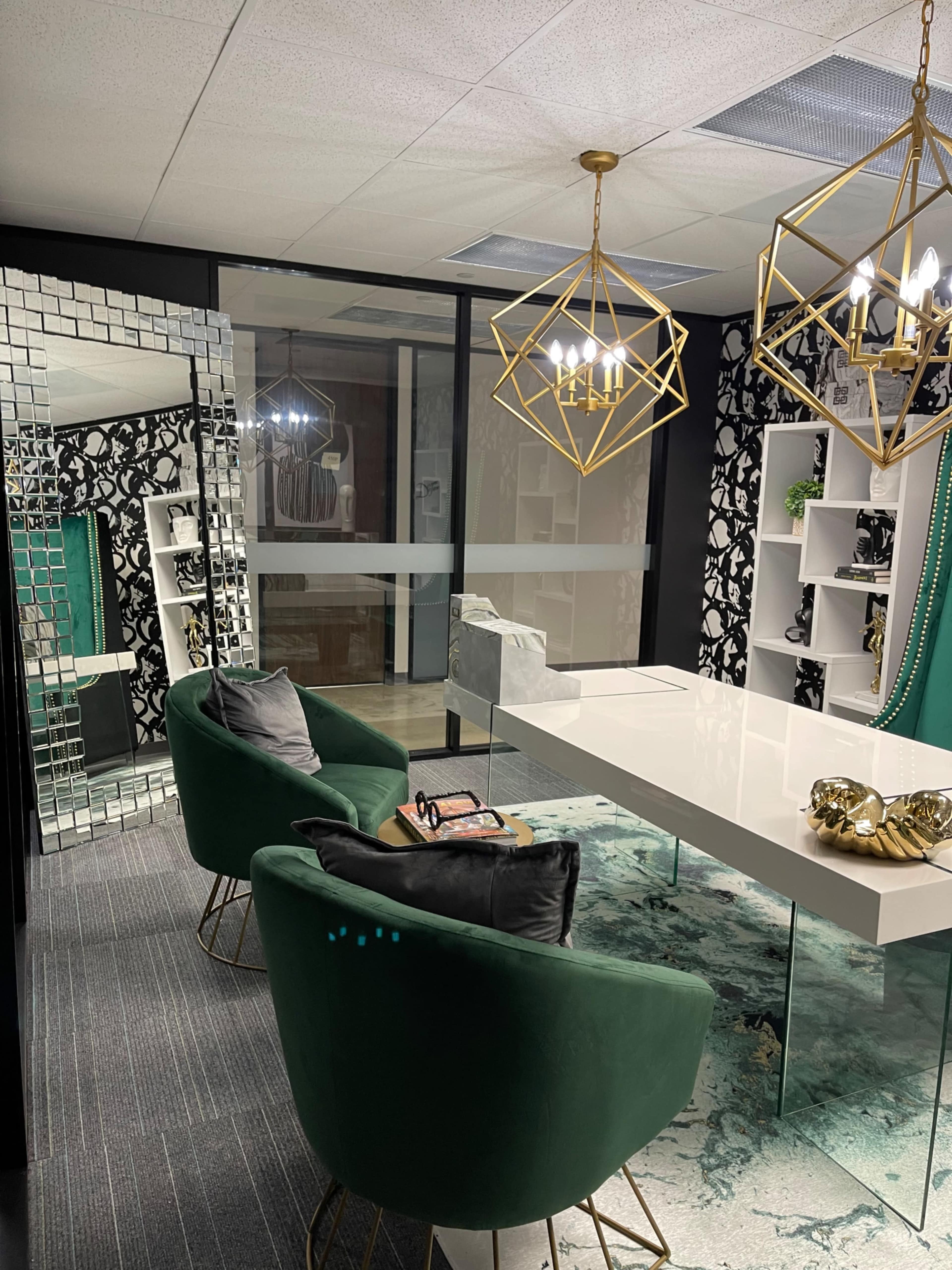 A modern office space featuring two green chairs, a white desk, decorative shelves, a decorative mirror, and geometric light fixtures against a patterned wallpaper backdrop.