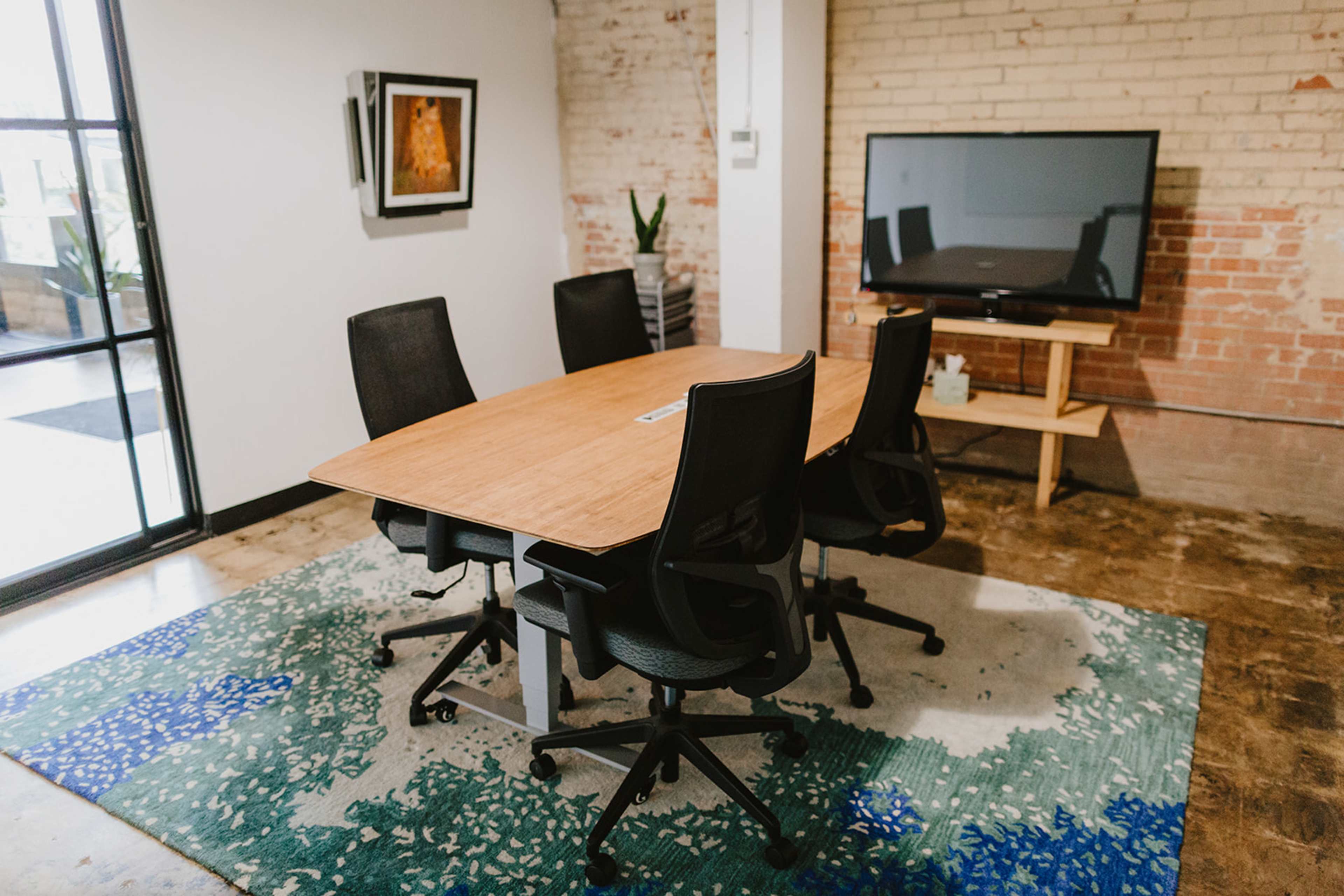 A conference room features a large wooden table surrounded by five black rolling chairs, with a television mounted on the wall and a patterned rug on the floor.