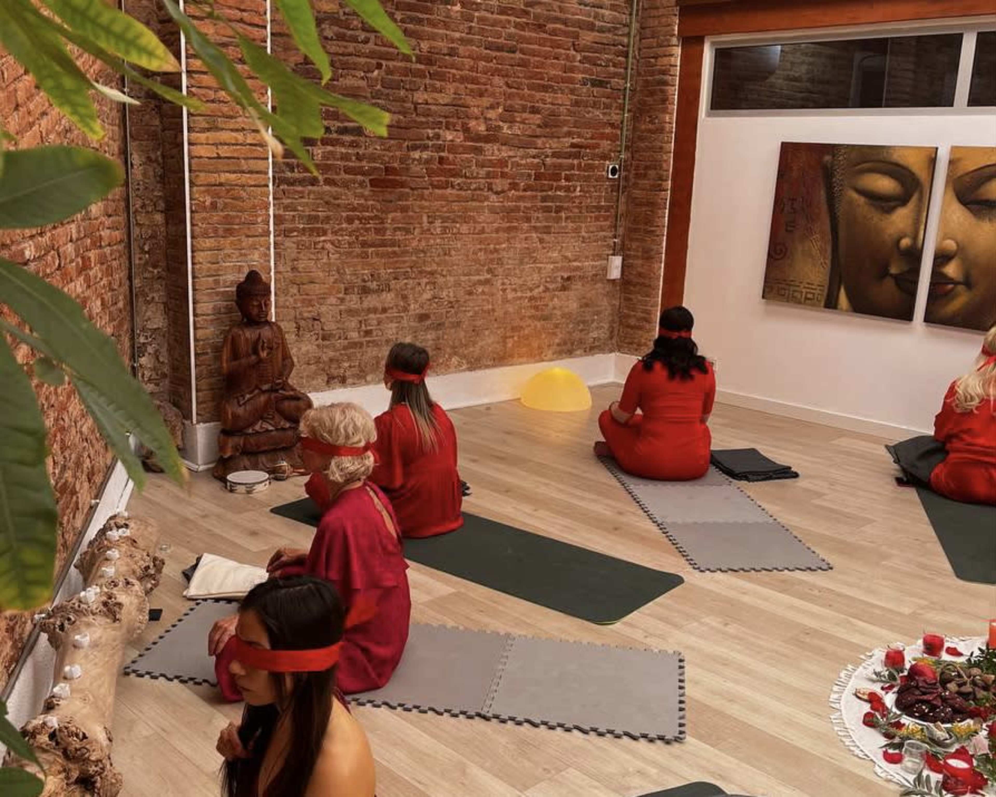 A group of individuals in red clothing sits on mats in a minimalist room with brick walls, facing a statue of Buddha.