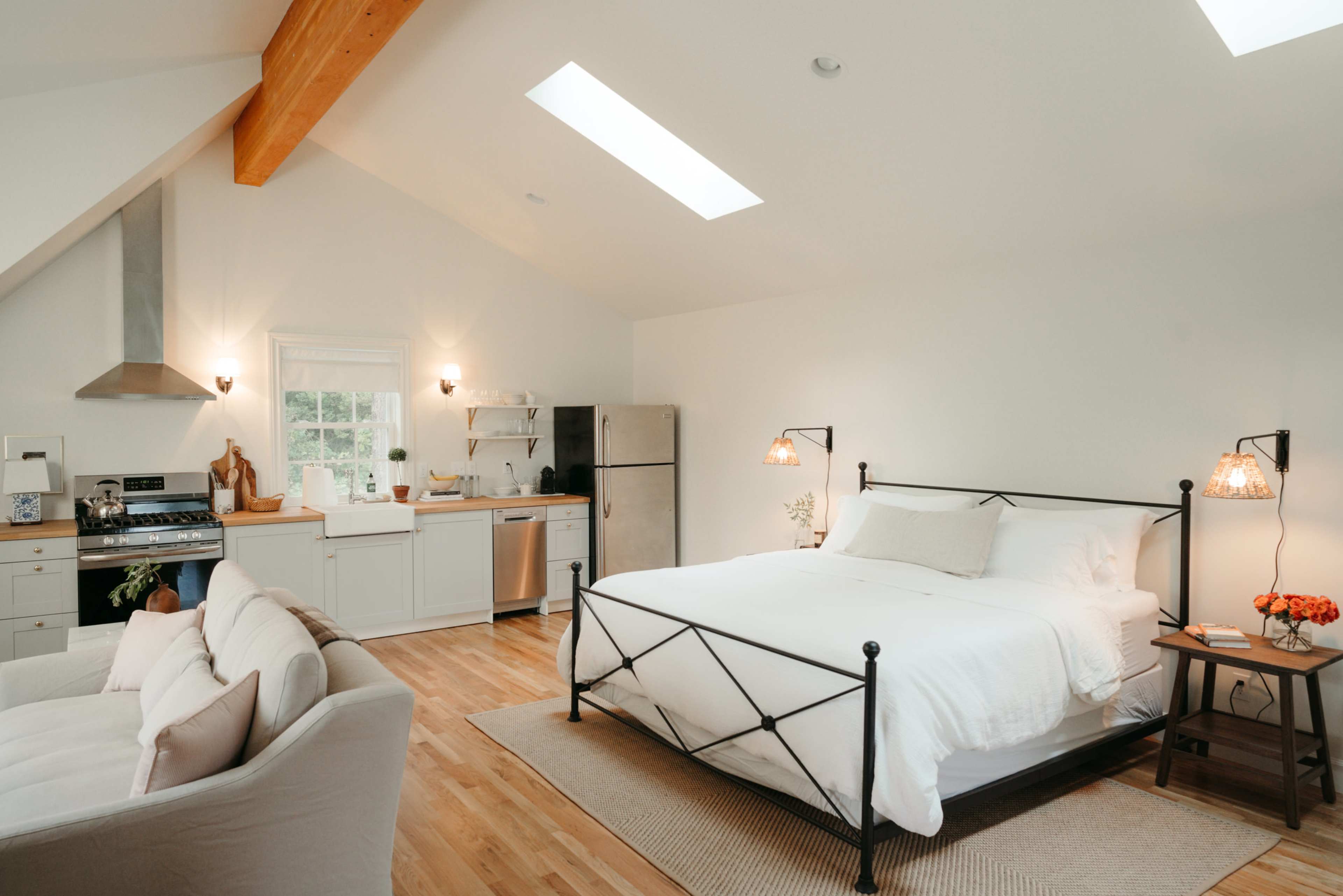 A cozy bedroom featuring a queen-sized bed with a black metal frame, accompanied by a kitchen area and a seating space with a light-colored sofa.