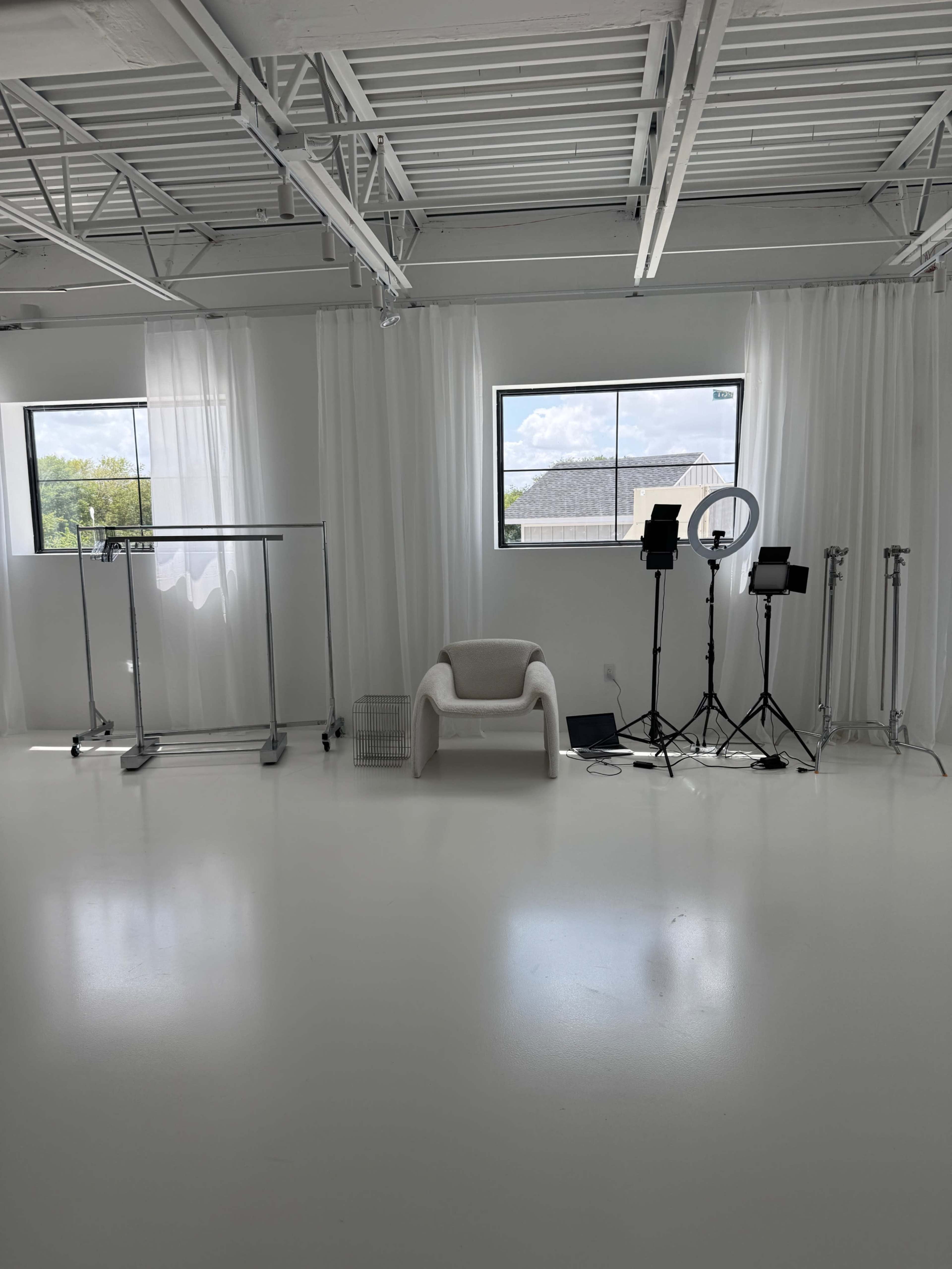 The image shows a bright, minimalist studio space with clothing racks, a white chair, and lighting equipment near large windows.