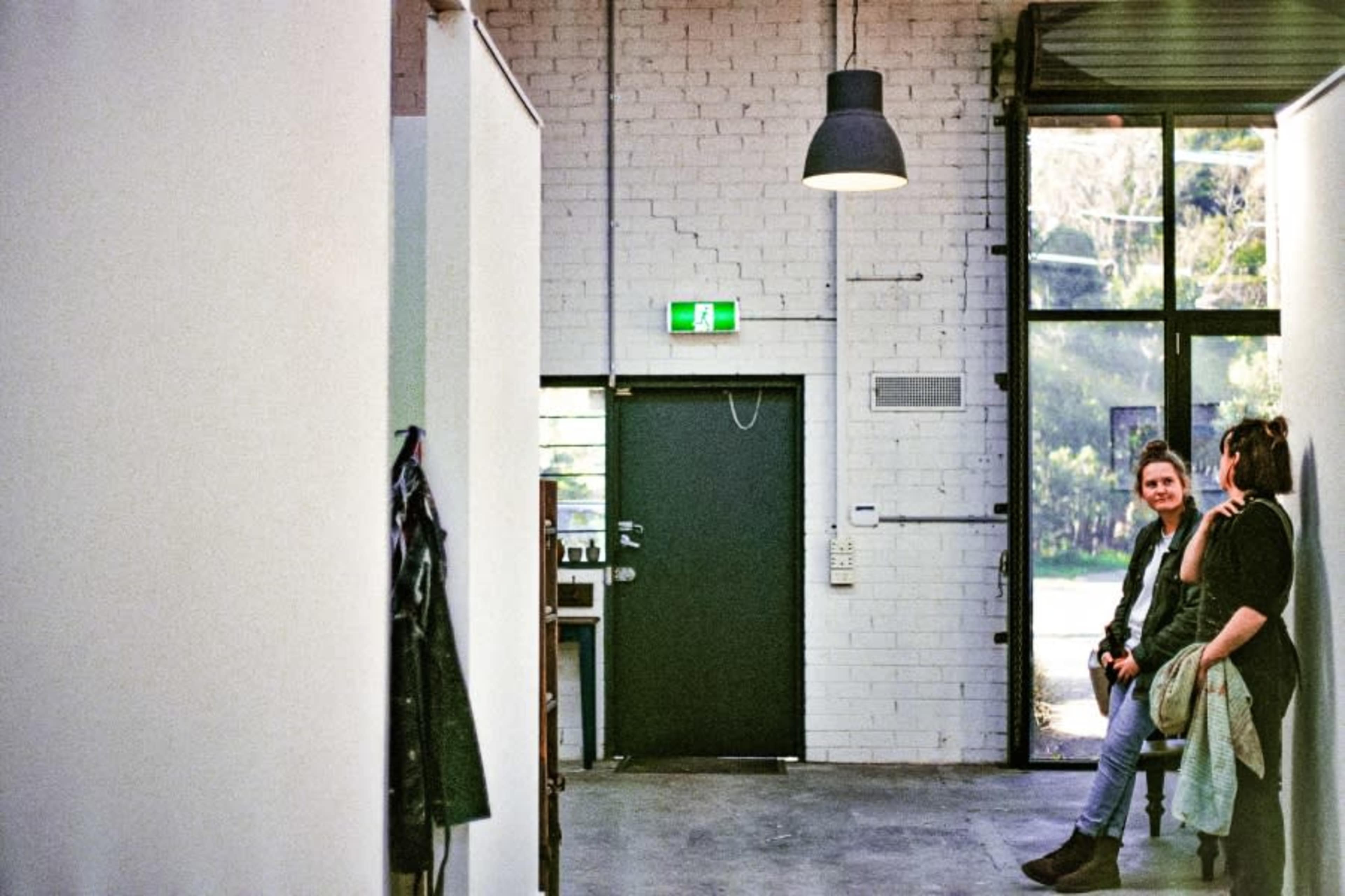 A woman sits on a ledge against a wall while another woman stands nearby in a spacious, brightly lit interior featuring white brick and large windows.