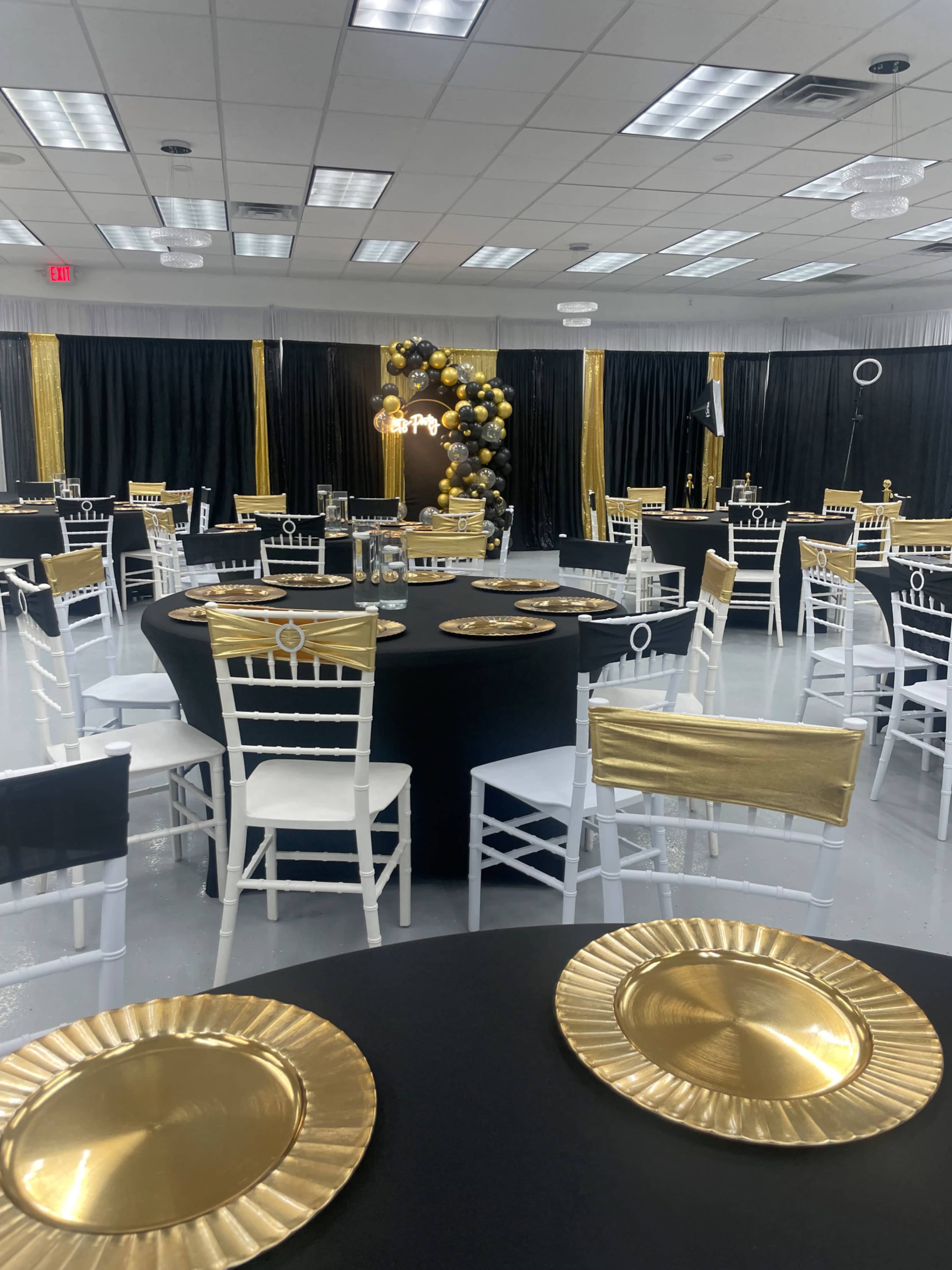 A banquet hall is set up with black and gold decor, featuring round black tables, white chairs, and decorative gold plates.