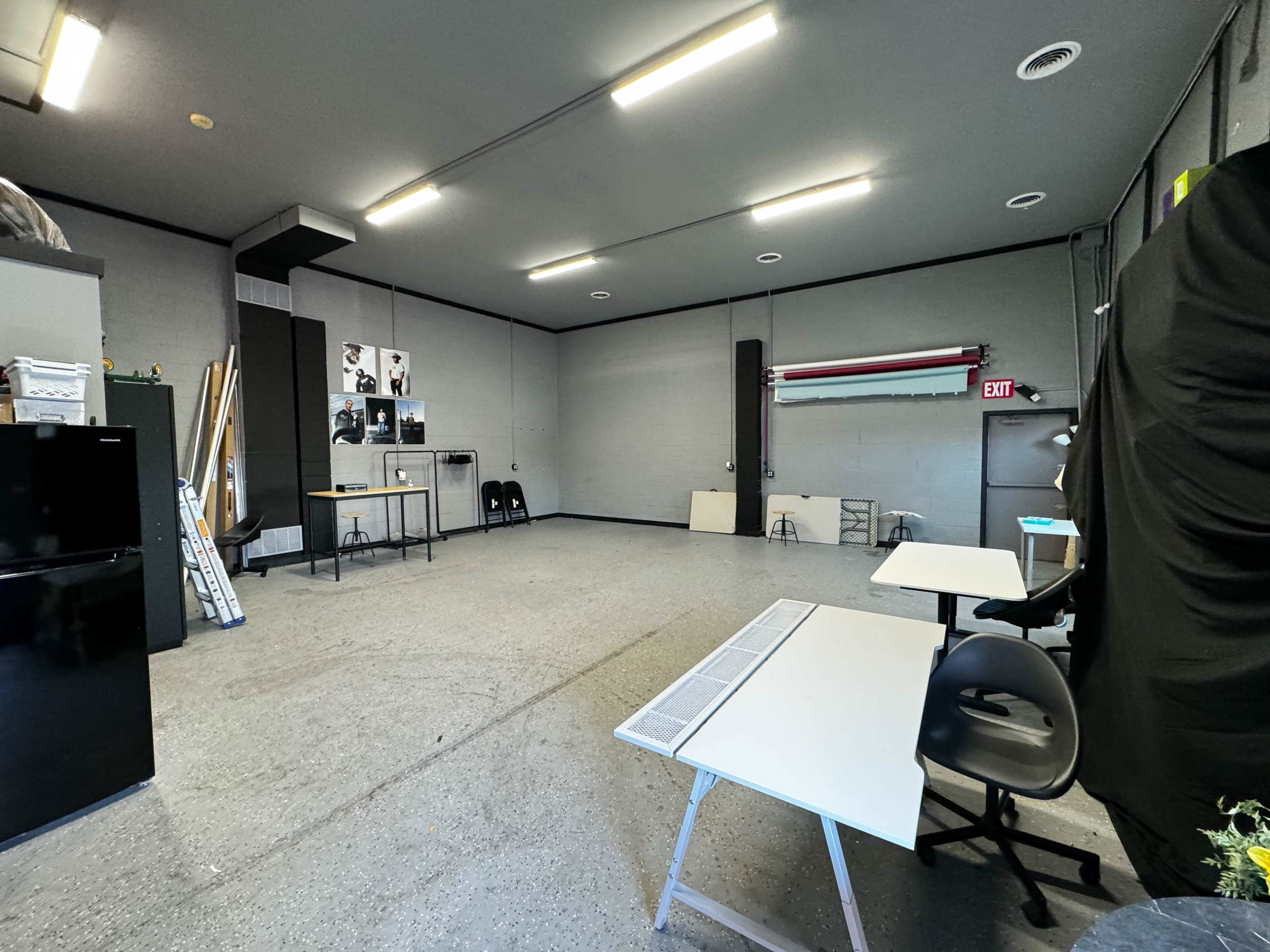 A spacious, empty room with grey walls, two tables, a black refrigerator, and light fixtures on the ceiling.