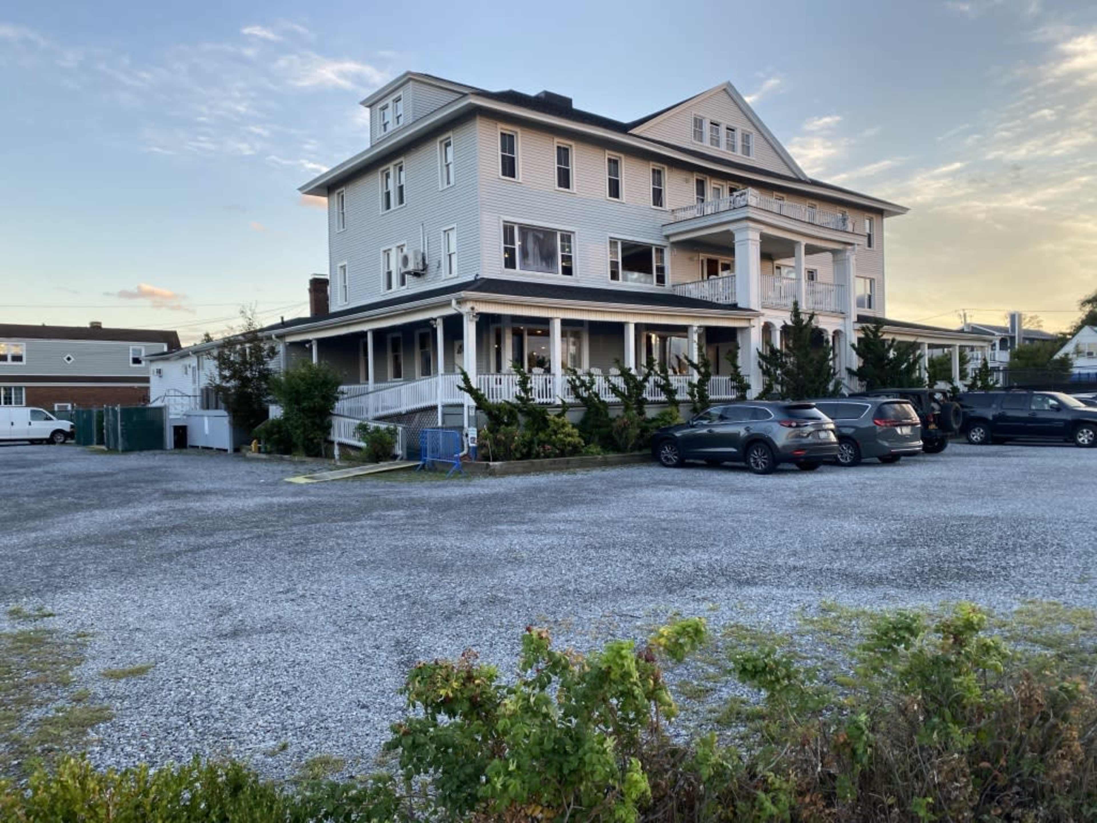 A large, multi-story building with a wraparound porch stands alongside a gravel parking lot filled with cars.