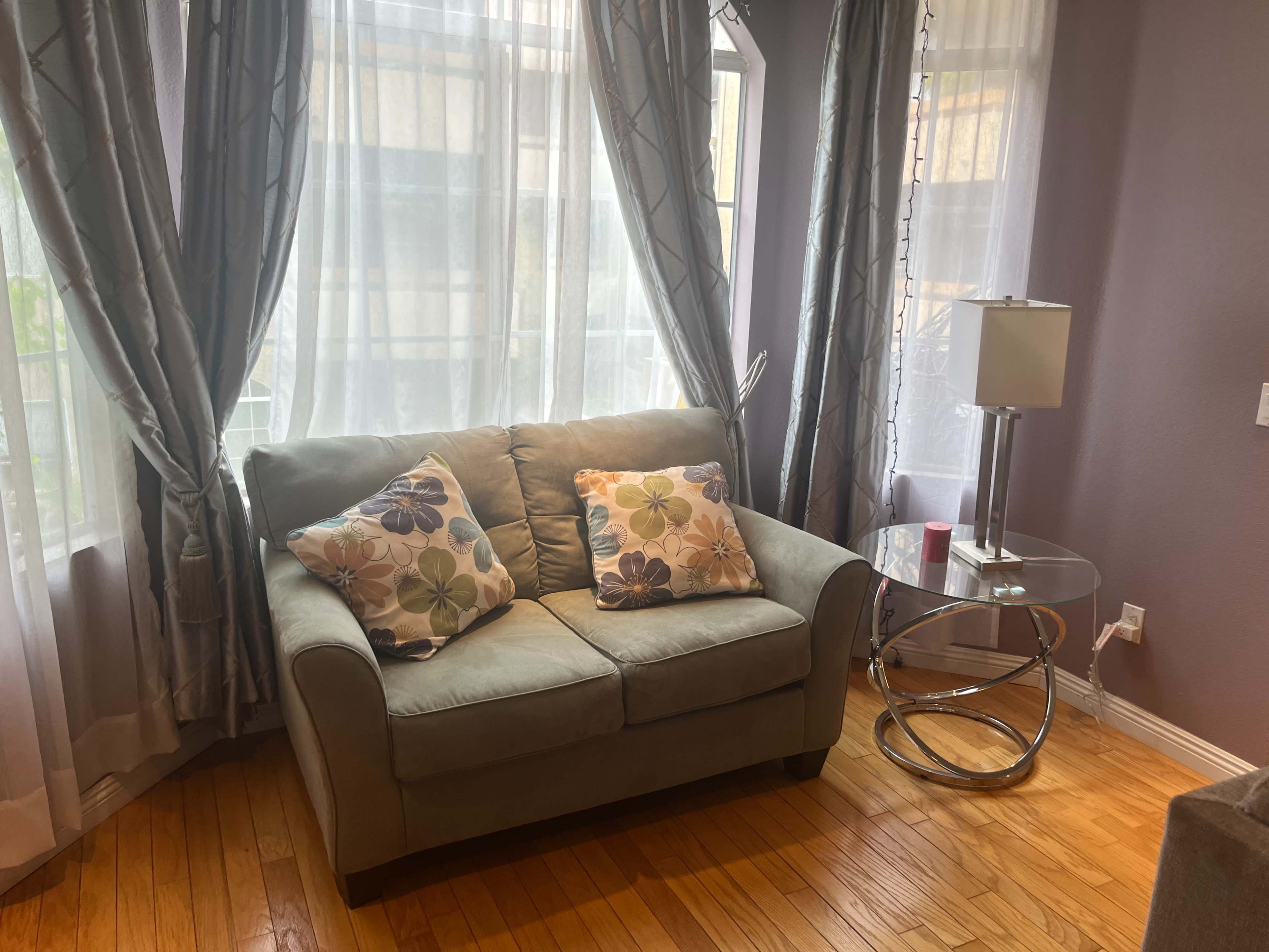 A light green loveseat with floral cushions is positioned beside a round glass side table near large, sheer curtains in a well-lit room.