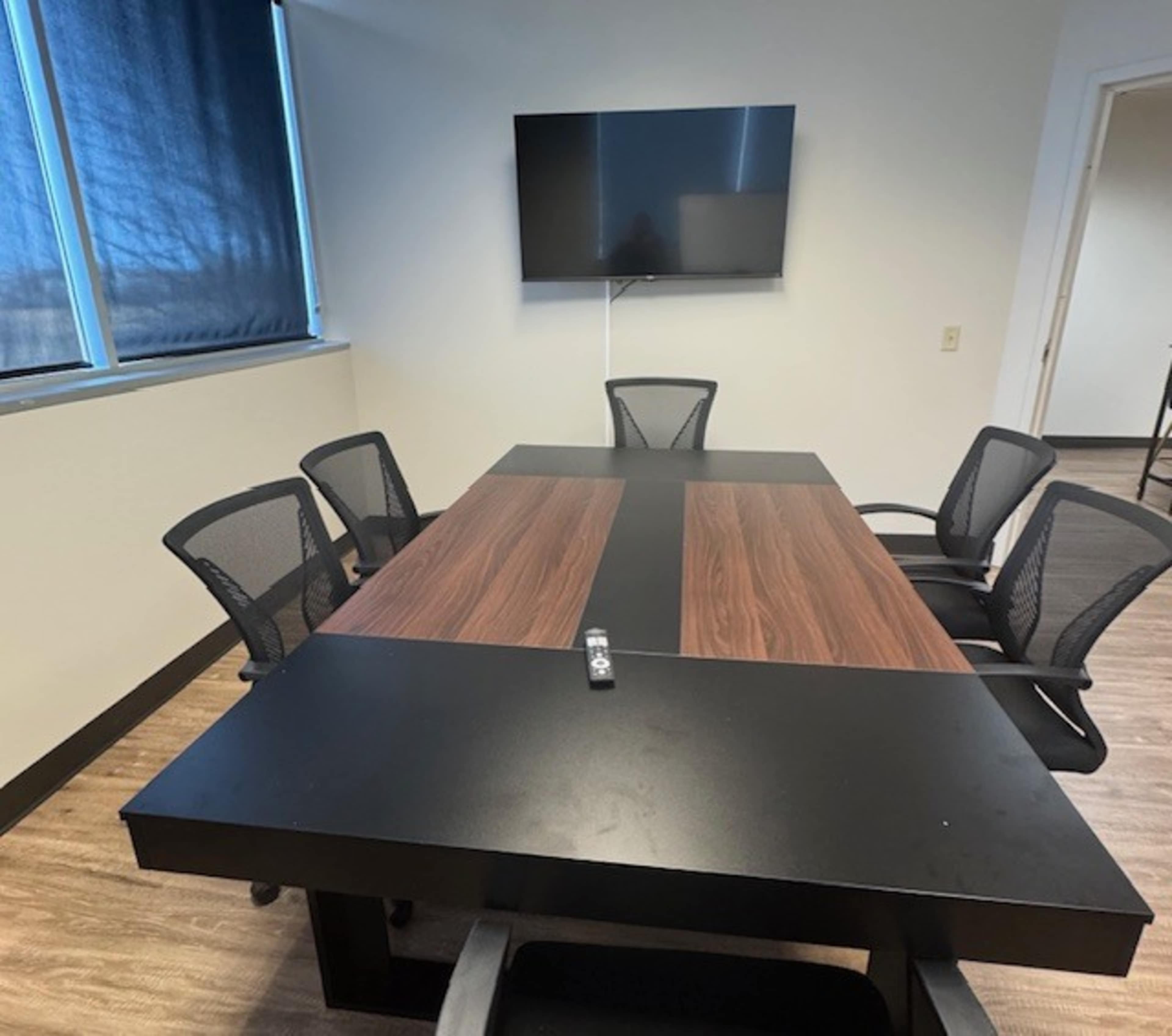 A modern conference room features a rectangular table with a wood finish, surrounded by black mesh chairs, and a large screen mounted on the wall.