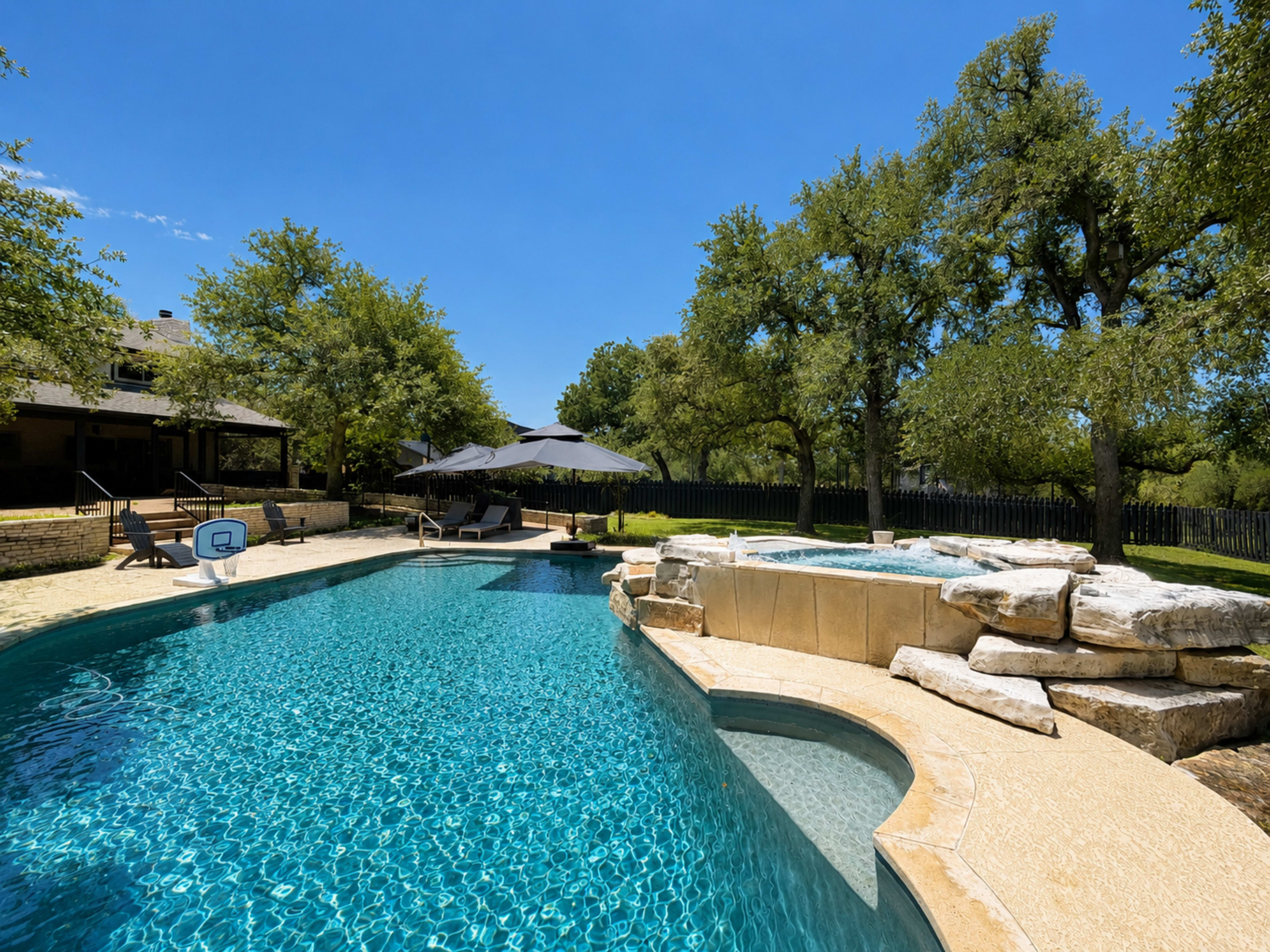 The image shows a swimming pool with a spa, surrounded by trees and outdoor seating areas, under a clear blue sky.