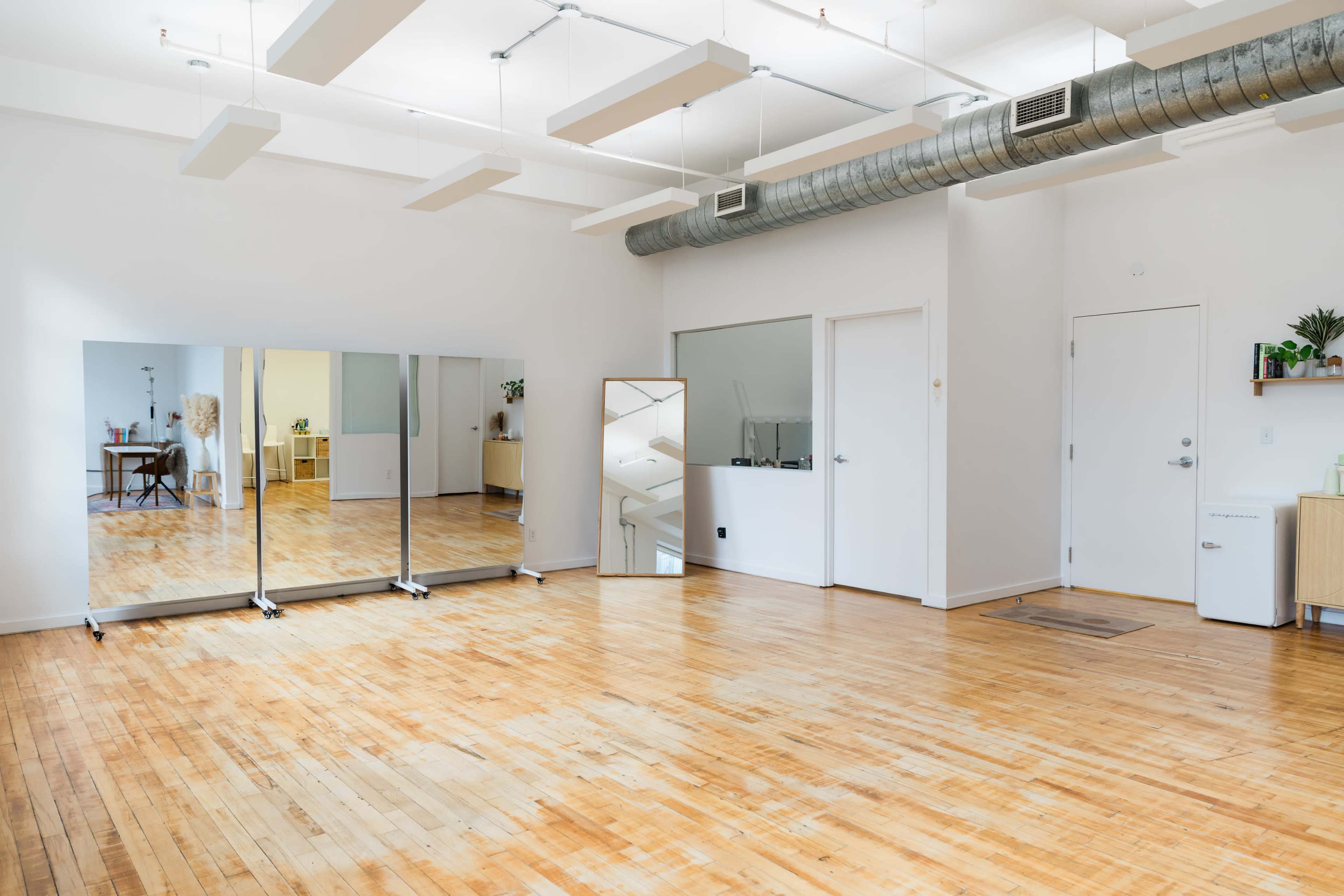 The image shows a bright, empty studio space with wooden flooring, large mirrors, and minimal furniture against white walls.