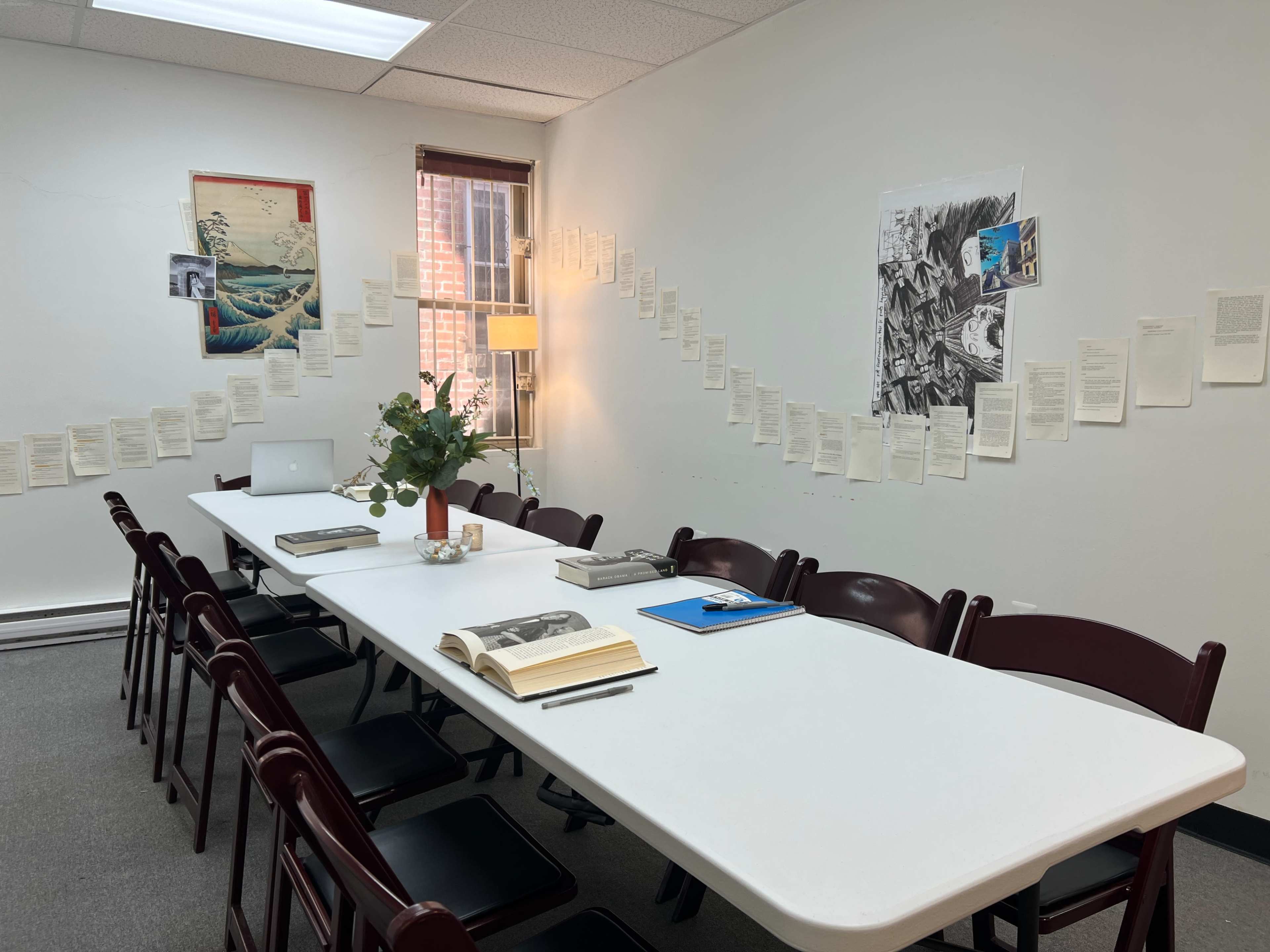 The image shows a meeting room with a long white table set with books, a laptop, and a floral centerpiece, surrounded by chairs, and walls adorned with pages of text and artwork.