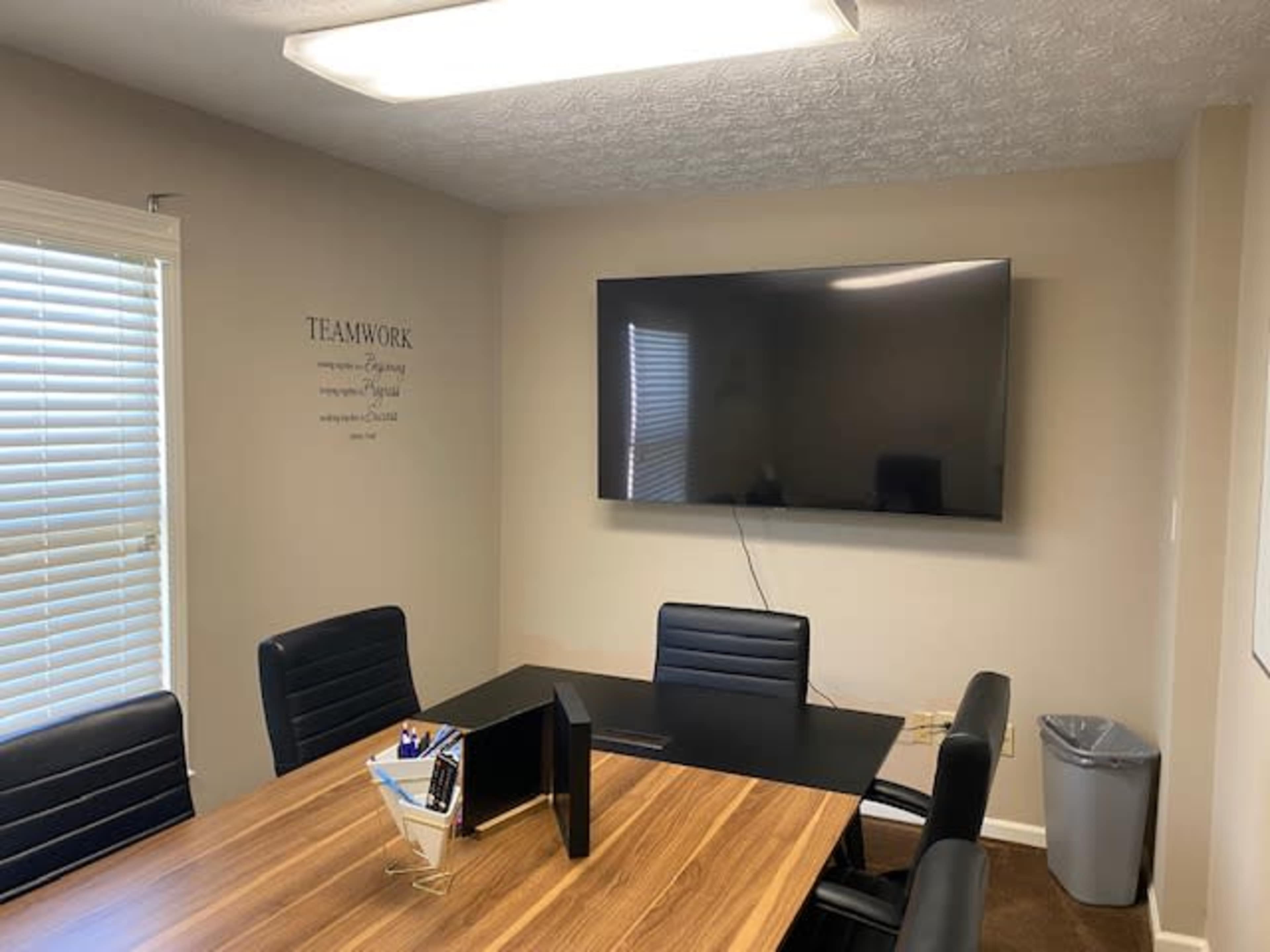 A conference room features a wooden table, black chairs, a large TV mounted on the wall, and a wastebasket in the corner.