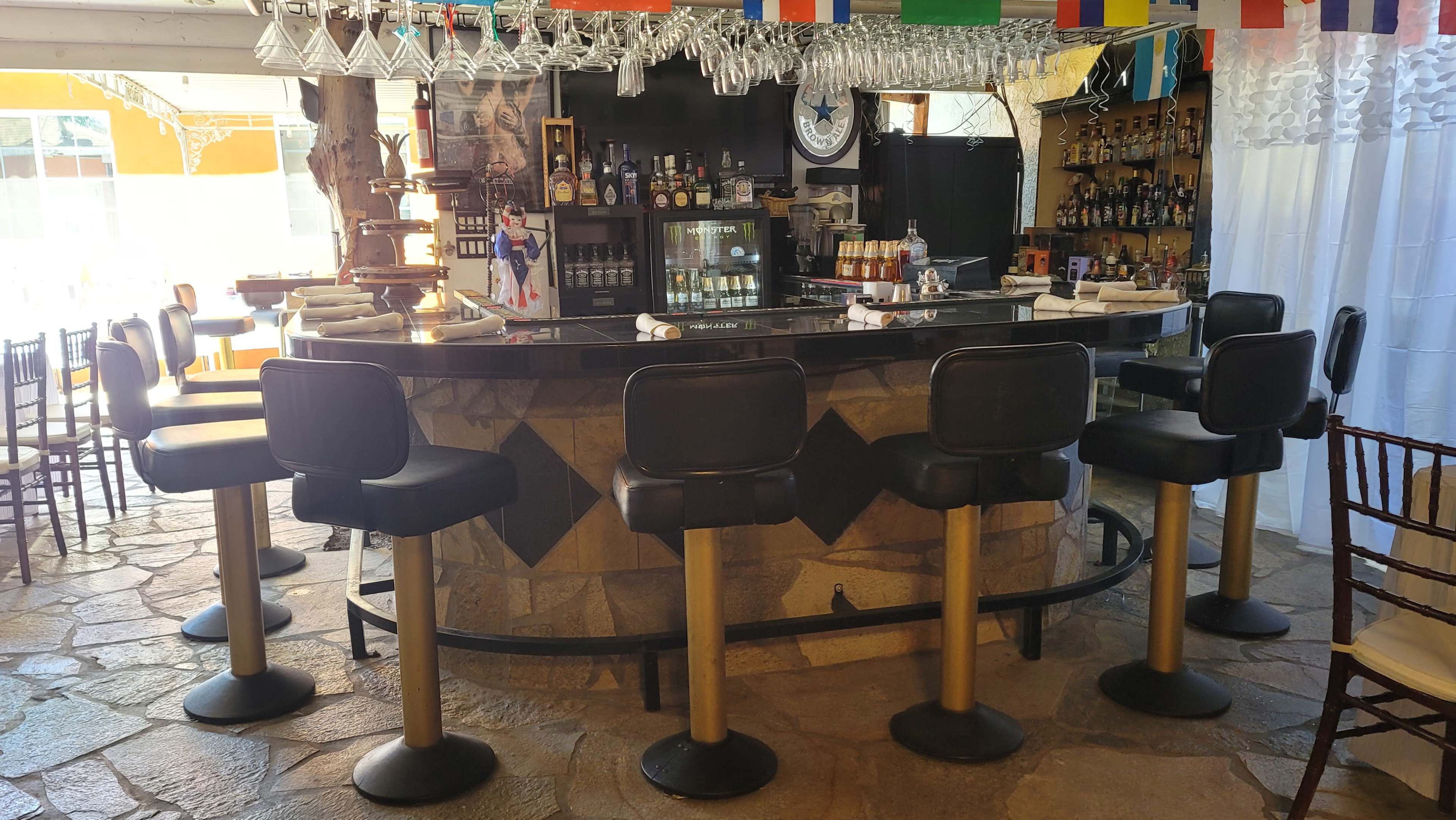 A bar with several black stools arranged around a stone countertop, surrounded by shelves stocked with various bottles and flags hanging overhead.