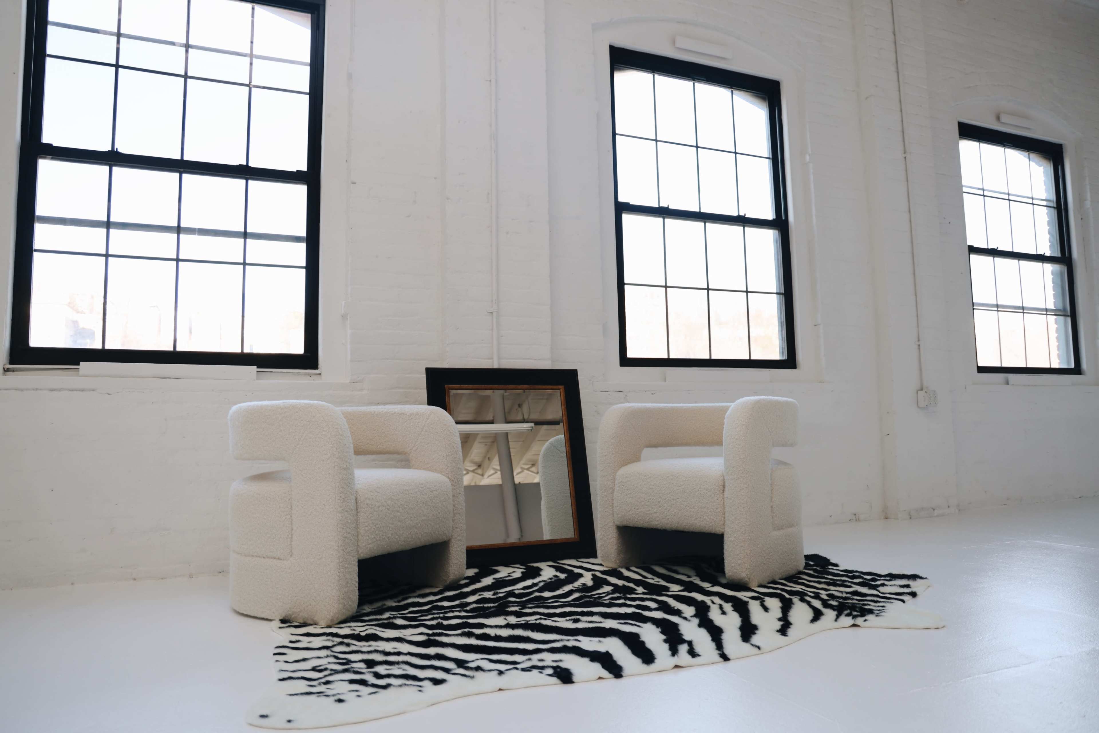 Two white upholstered chairs are positioned on a zebra print rug in a bright room with large windows and a mirror leaning against the wall.