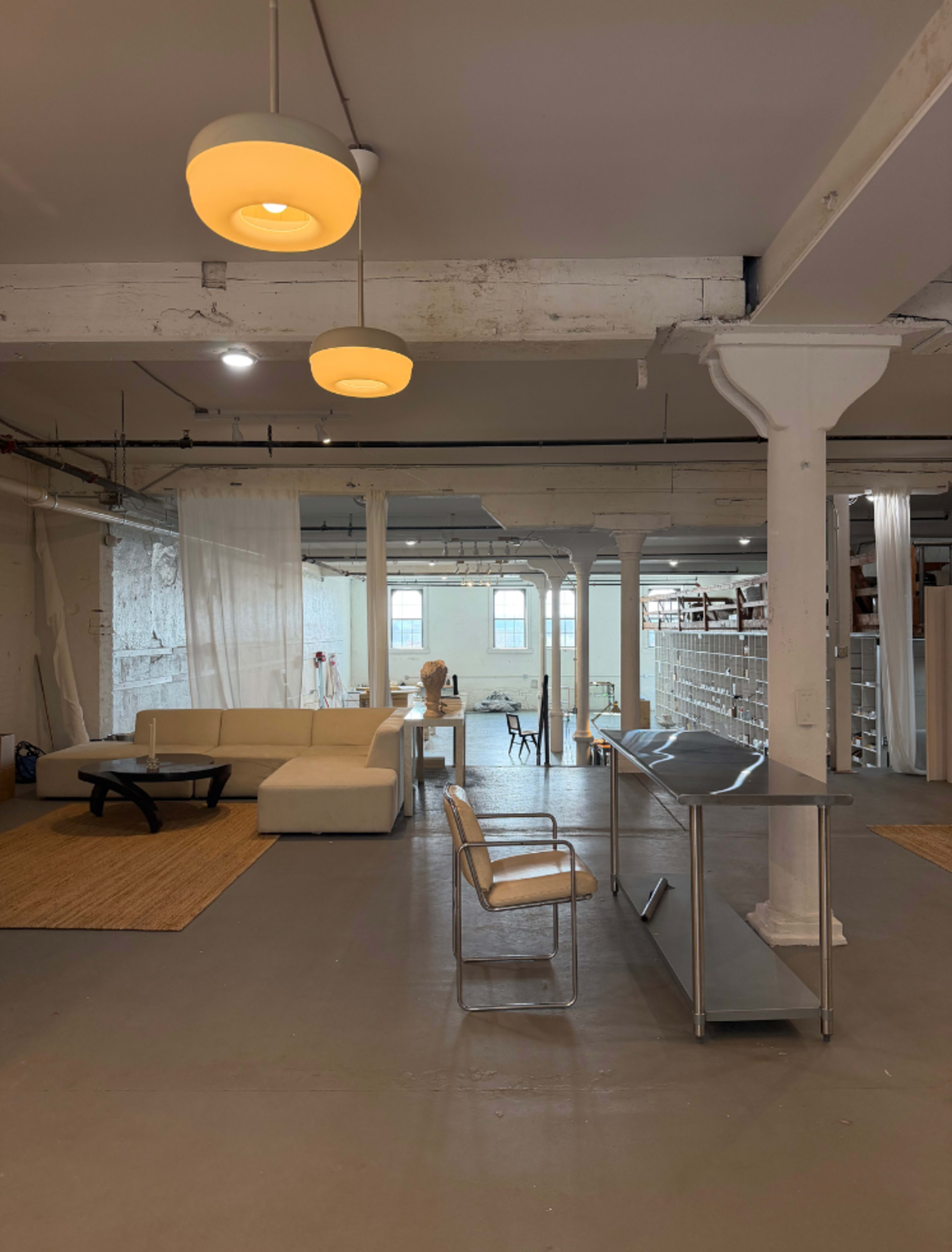 The image shows a spacious, modern studio with a white couch, a glass-top table, and shelves filled with items in a well-lit environment.