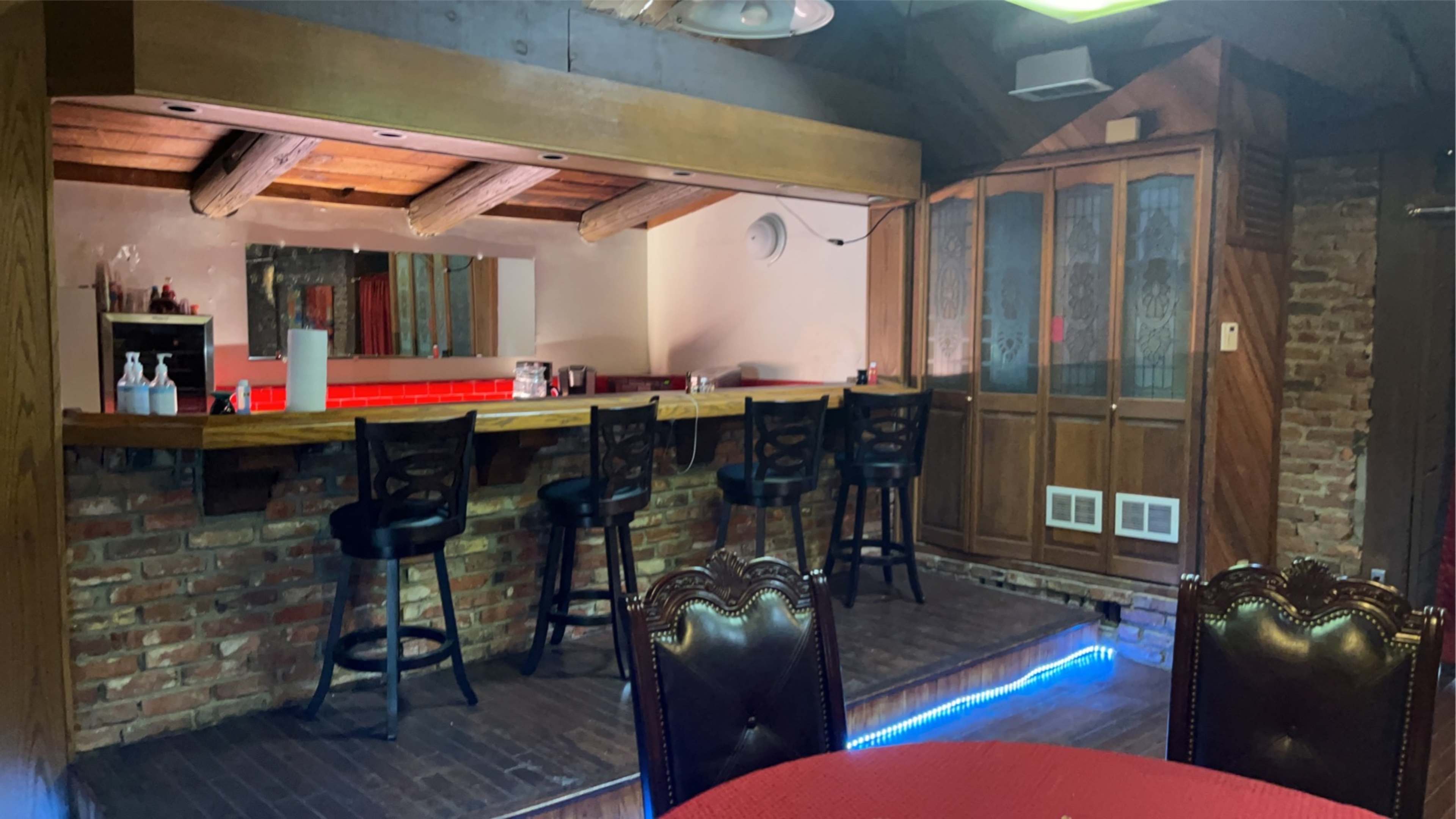 A bar area with a wooden counter and high black stools, set against a backdrop of exposed brick walls and wooden beams.