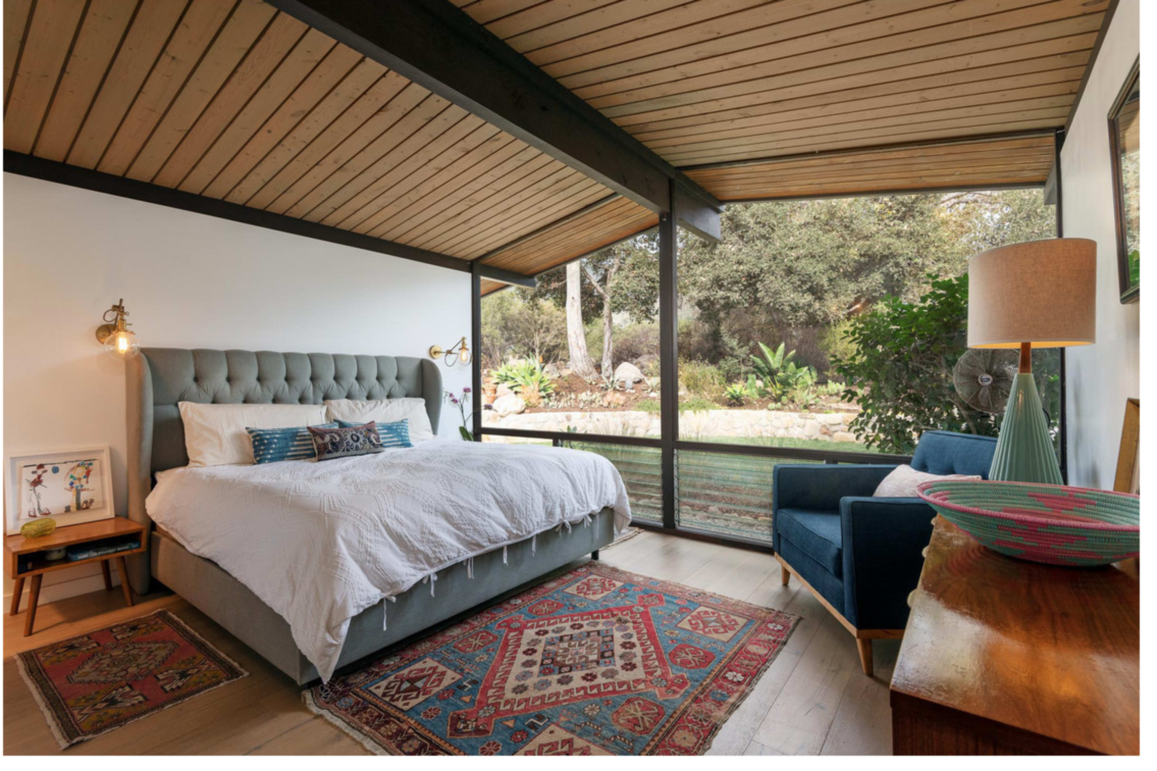 A modern bedroom features a tufted bed, a blue armchair, and large glass windows overlooking a garden.
