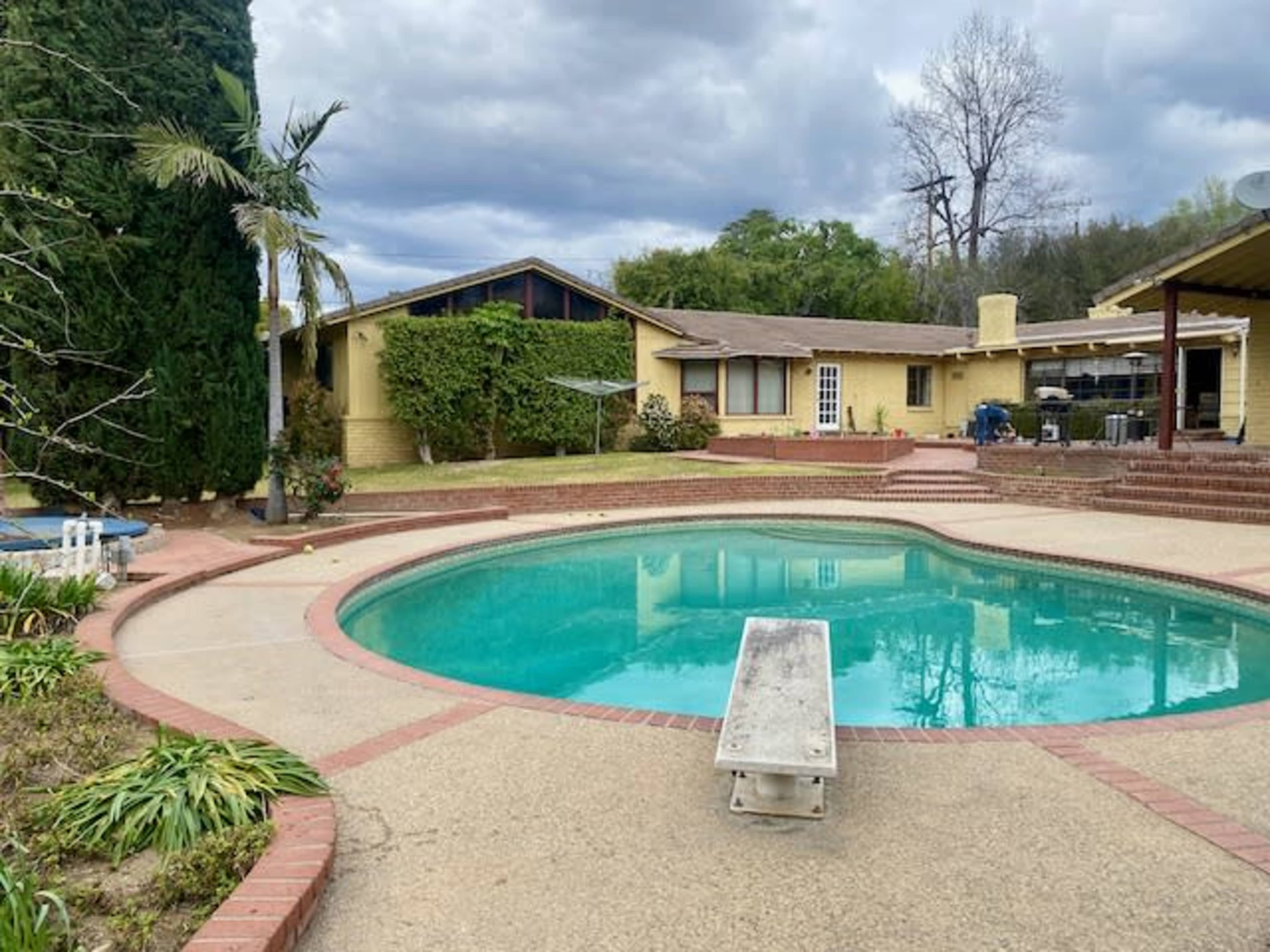 A circular swimming pool with a diving board is situated in a backyard surrounded by a single-story house and landscaped greenery under a cloudy sky.
