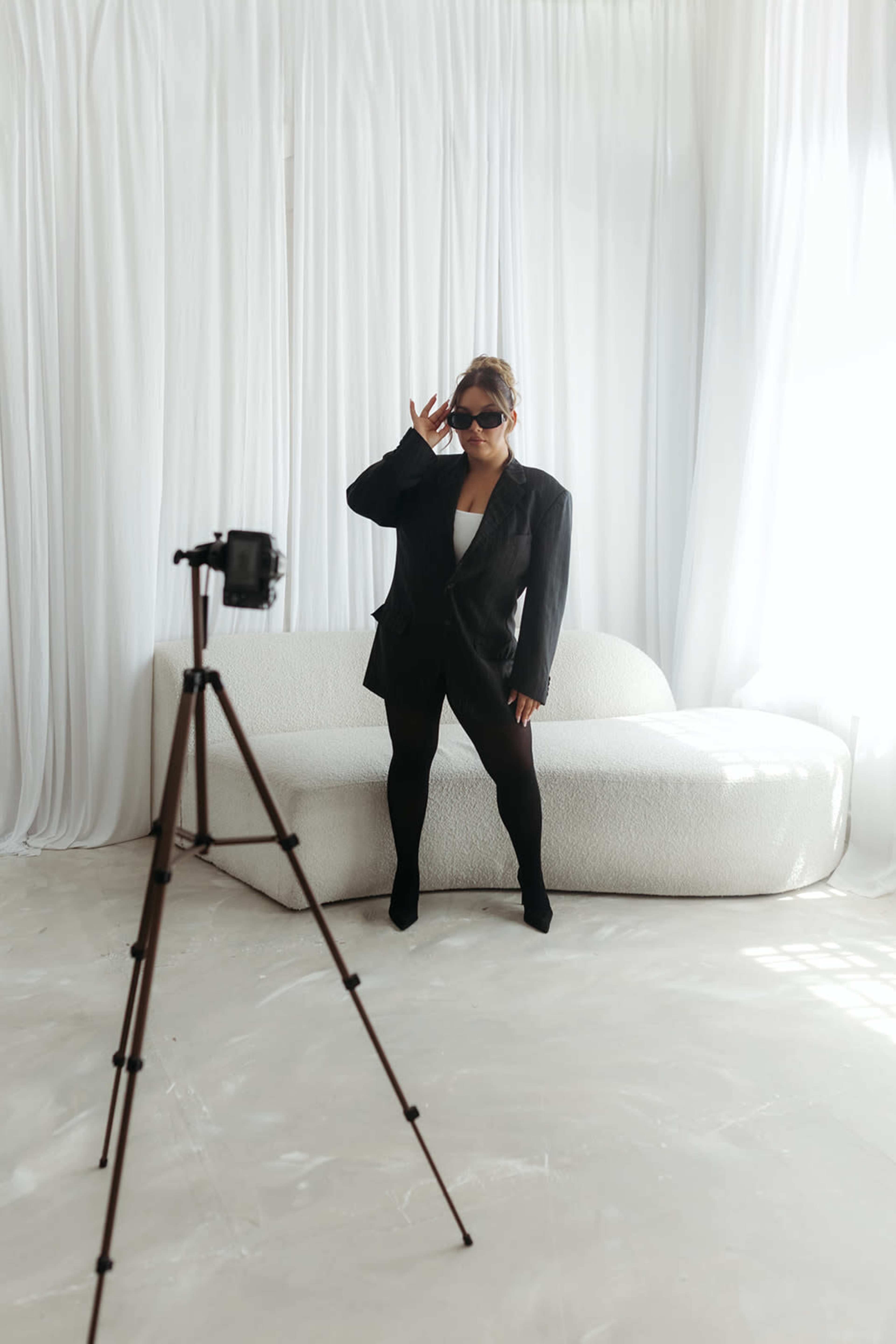 A person wearing a black blazer and sunglasses poses in front of a camera mounted on a tripod, set against a backdrop of white drapes and a light-colored couch.