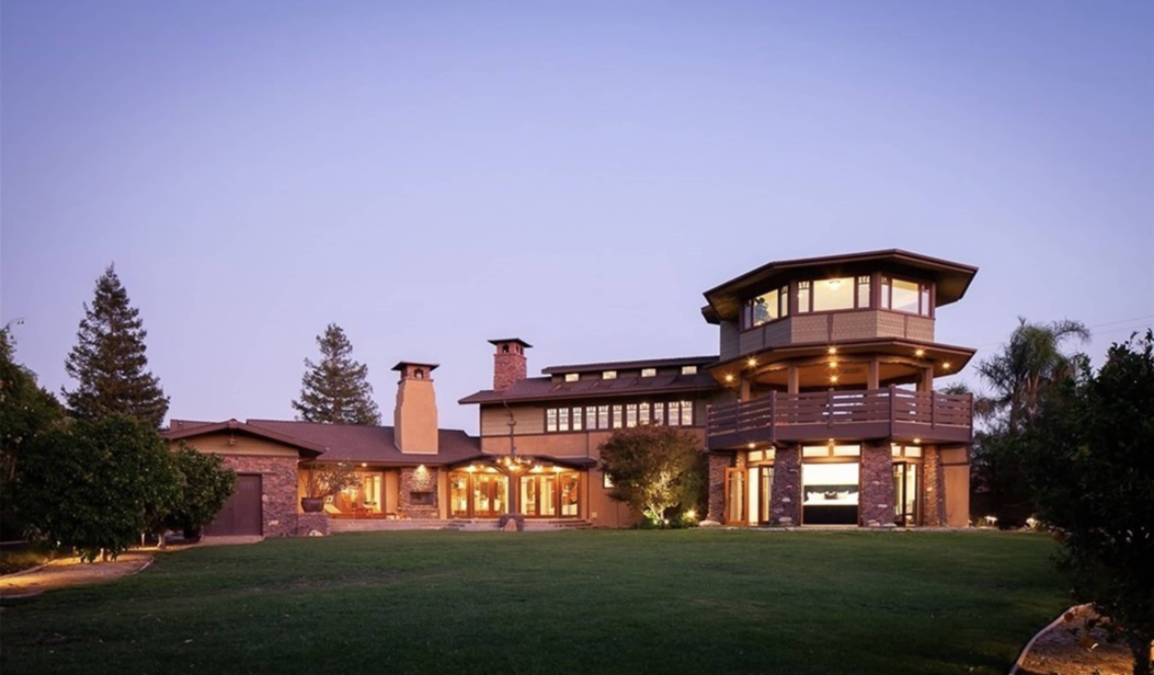 The image shows a multi-level house with a tower and large windows, surrounded by a green lawn and trees in the evening light.