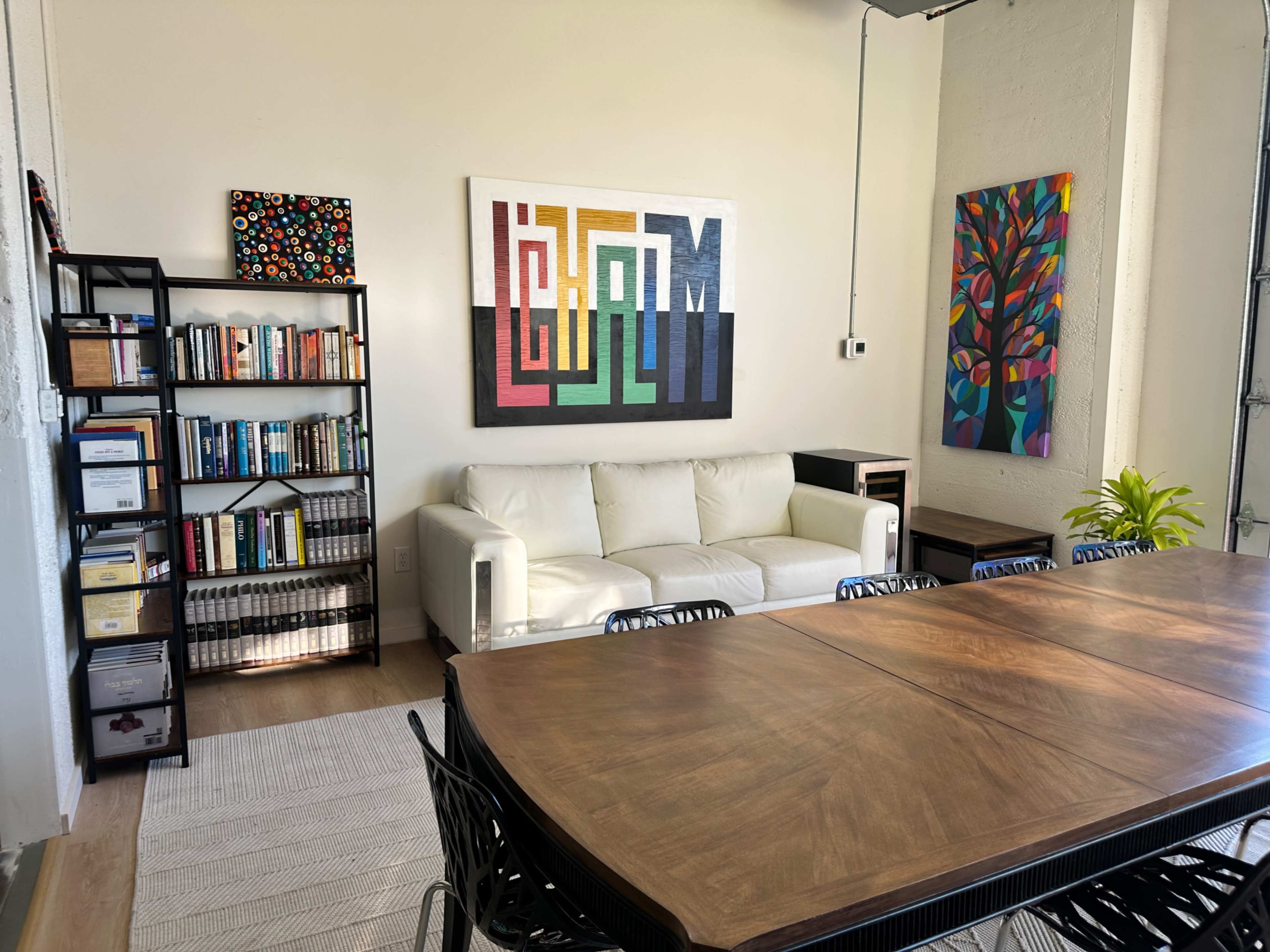 The image shows a bright, modern office space featuring a white couch, a wooden conference table, bookshelves filled with books and documents, and colorful artwork on the walls.