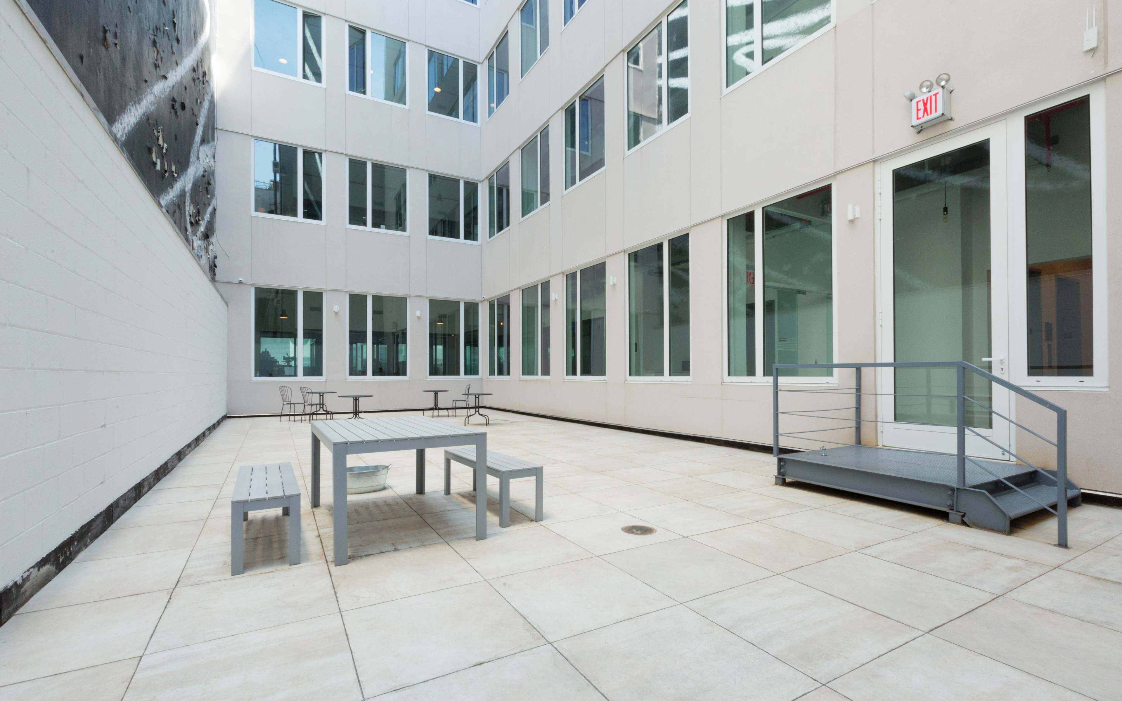 A modern outdoor courtyard with multiple tables and chairs, surrounded by glass walls.
