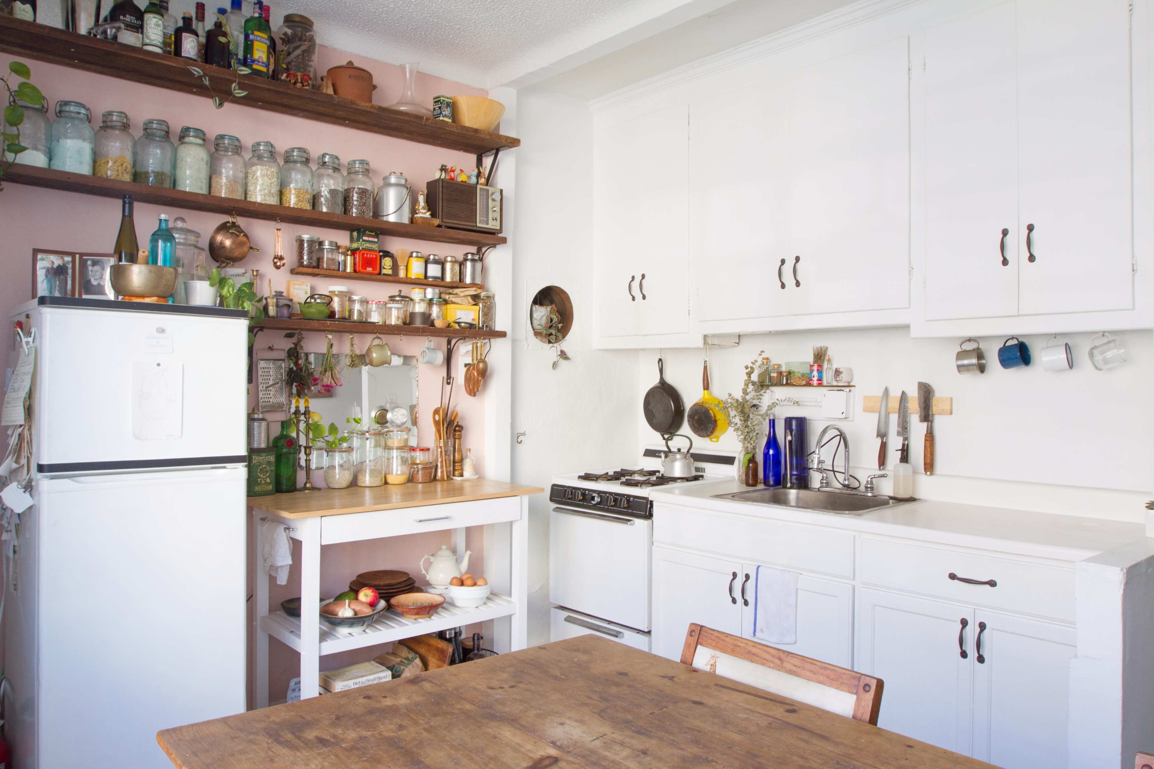 A bright kitchen with white cabinets, open shelving filled with jars and kitchen items, a stove, and a wooden table set for a meal.